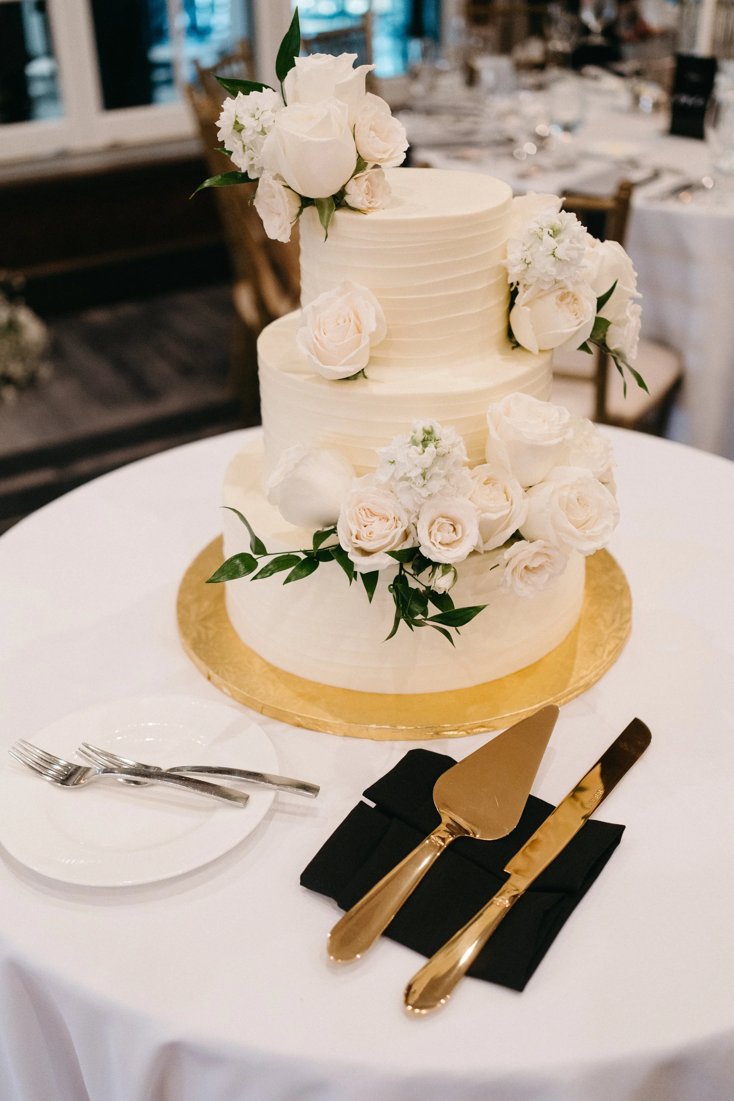 A two-tier white wedding cake decorated with white roses and greenery on top, placed on a table with a gold cake board. In front of the cake, there are gold and black utensils wrapped in a black napkin, set on a white tablecloth.