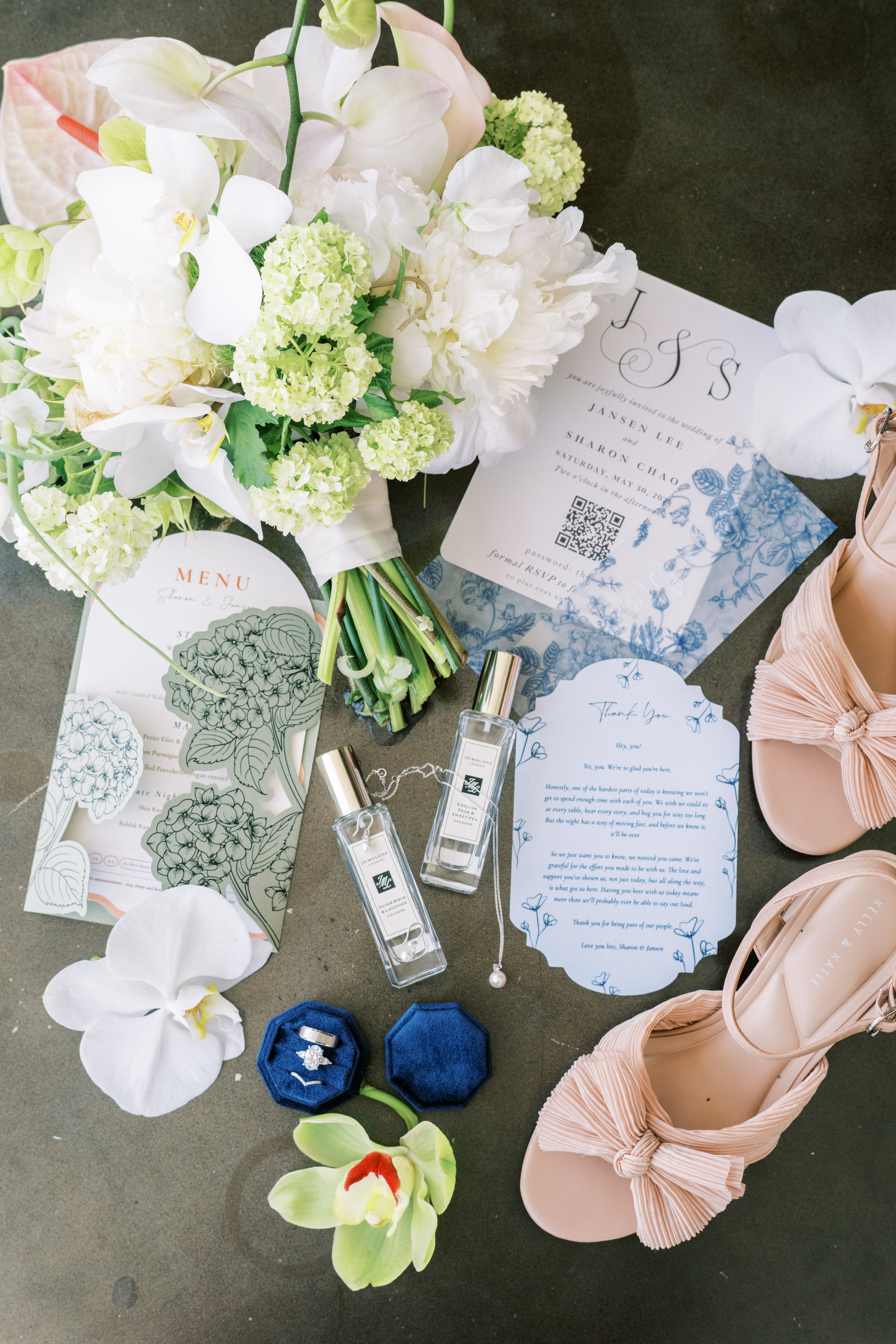 A wedding bouquet with white orchids and hydrangeas, wedding invitation and menu cards, two blue rings in a velvet box, a Thank You card, small perfume bottles, and pink high-heeled shoes with bows on a dark surface.
