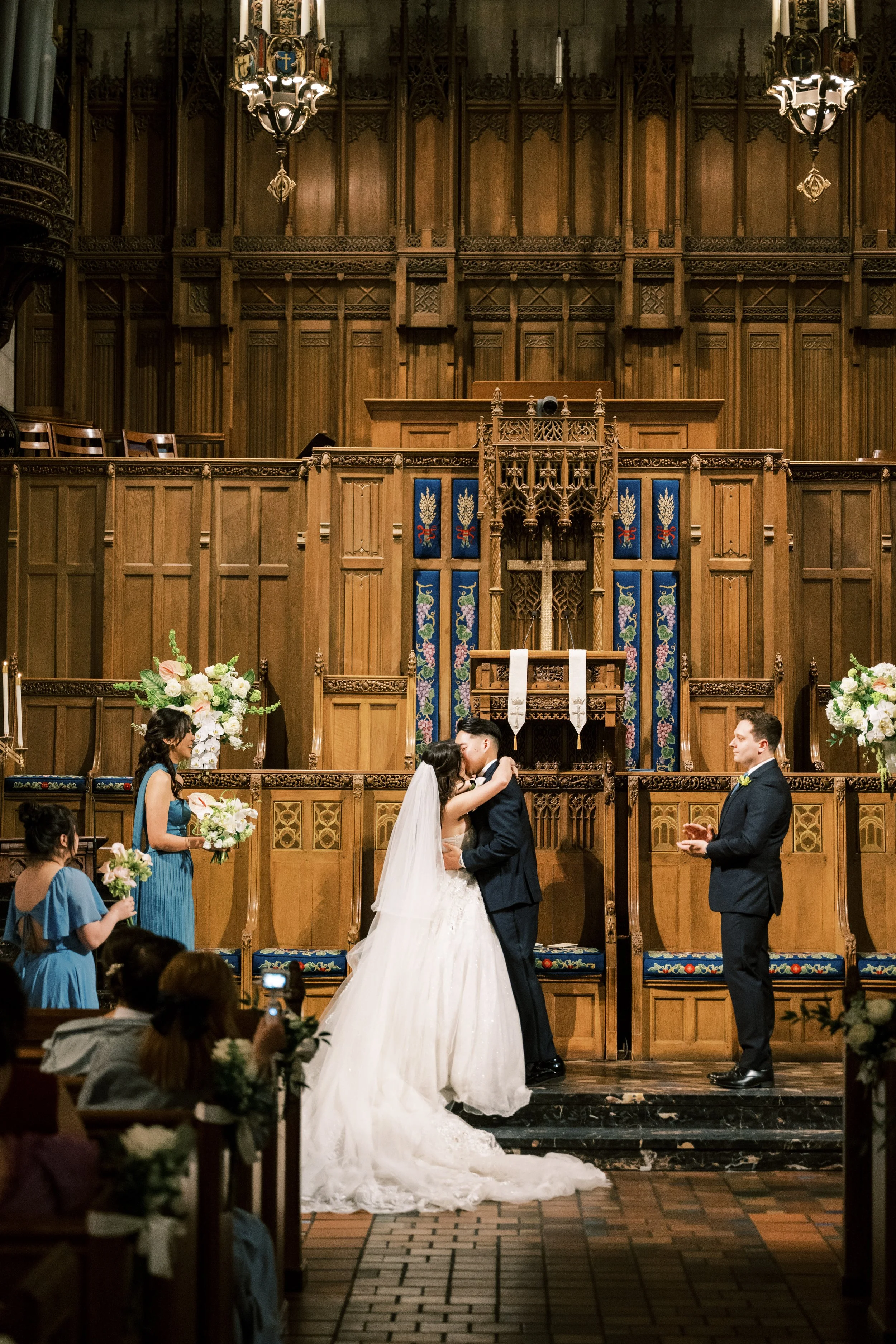 A wedding ceremony taking place in a church with a wooden altar and gothic architecture. The bride and groom are kissing at the altar, surrounded by bridesmaids and groomsmen. The bride wears a white wedding gown, and the groom wears a dark suit. Flo