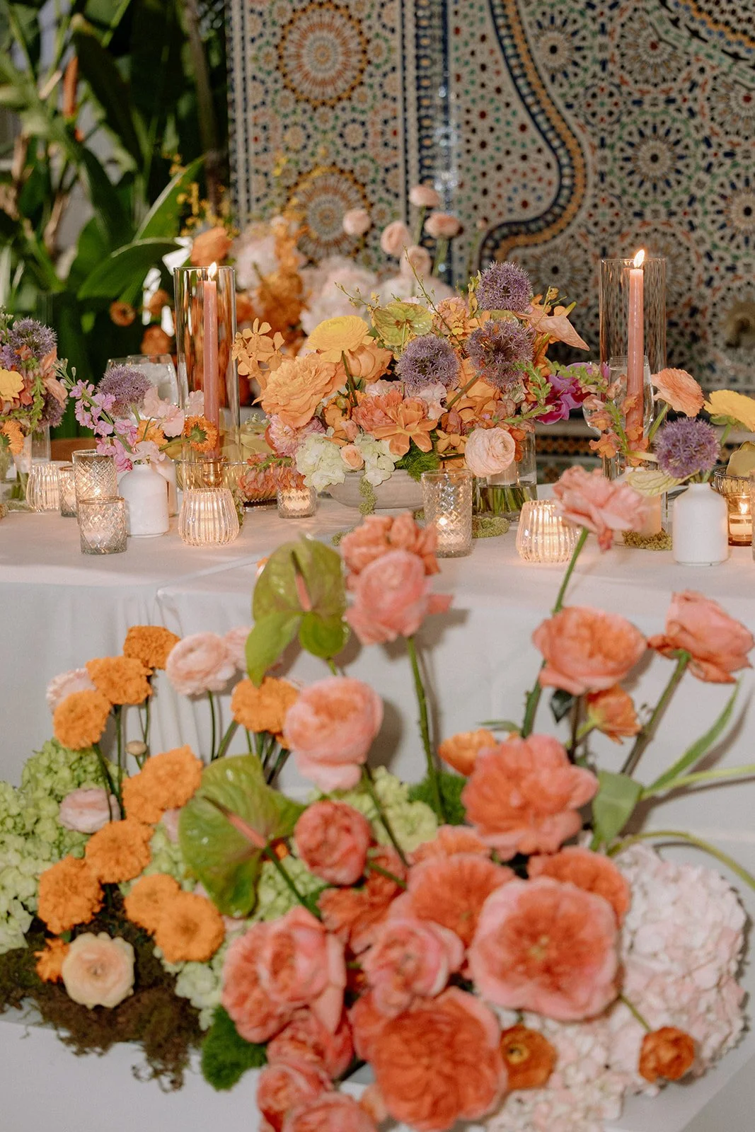 A table decorated with pink, purple, and orange flowers, surrounded by candles and glass vases, with a patterned wall in the background.