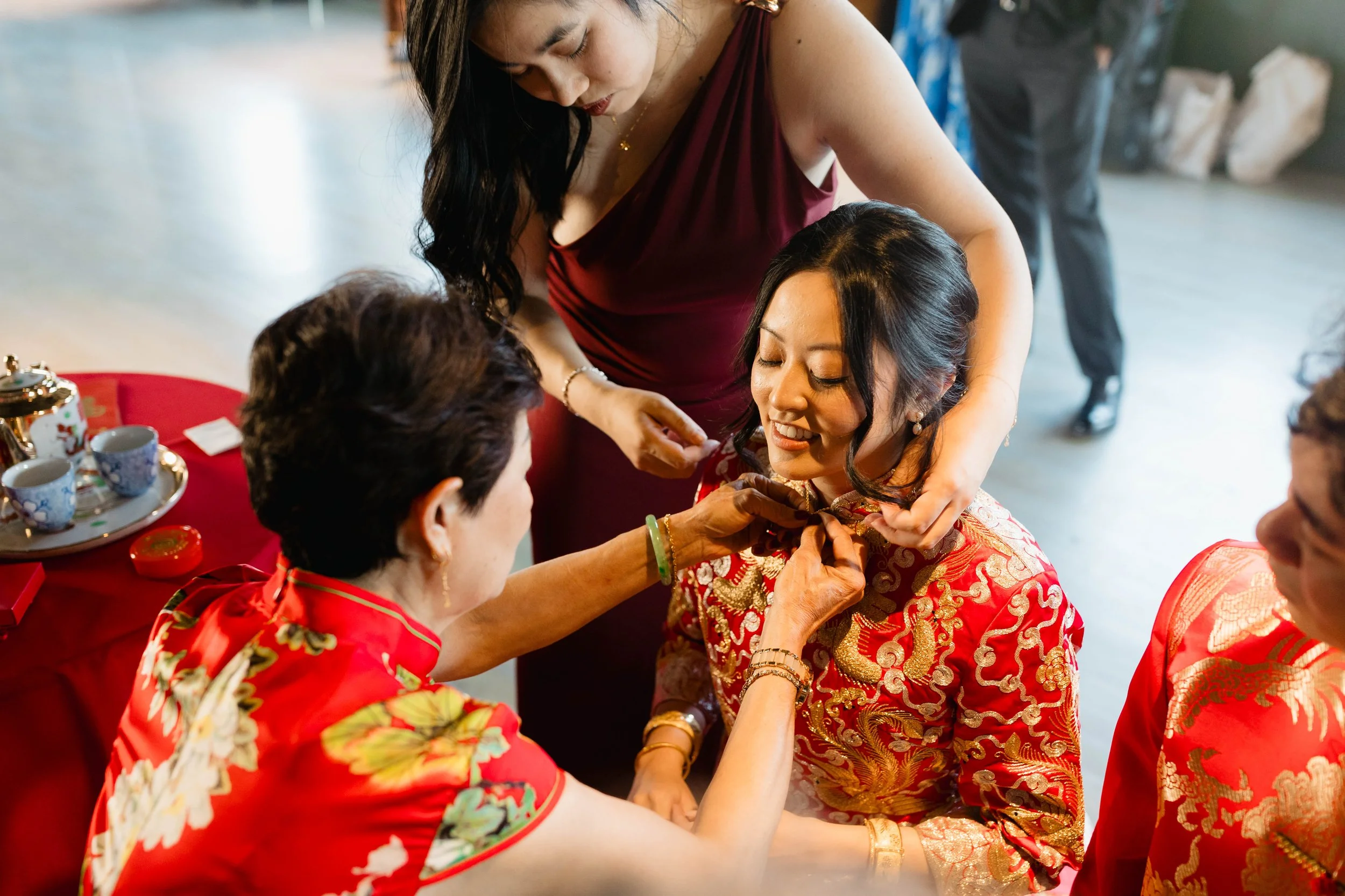 Women dressed in traditional red and gold Asian attire participate in a cultural ceremony, with one woman smiling while a hand places a bracelet on her wrist.