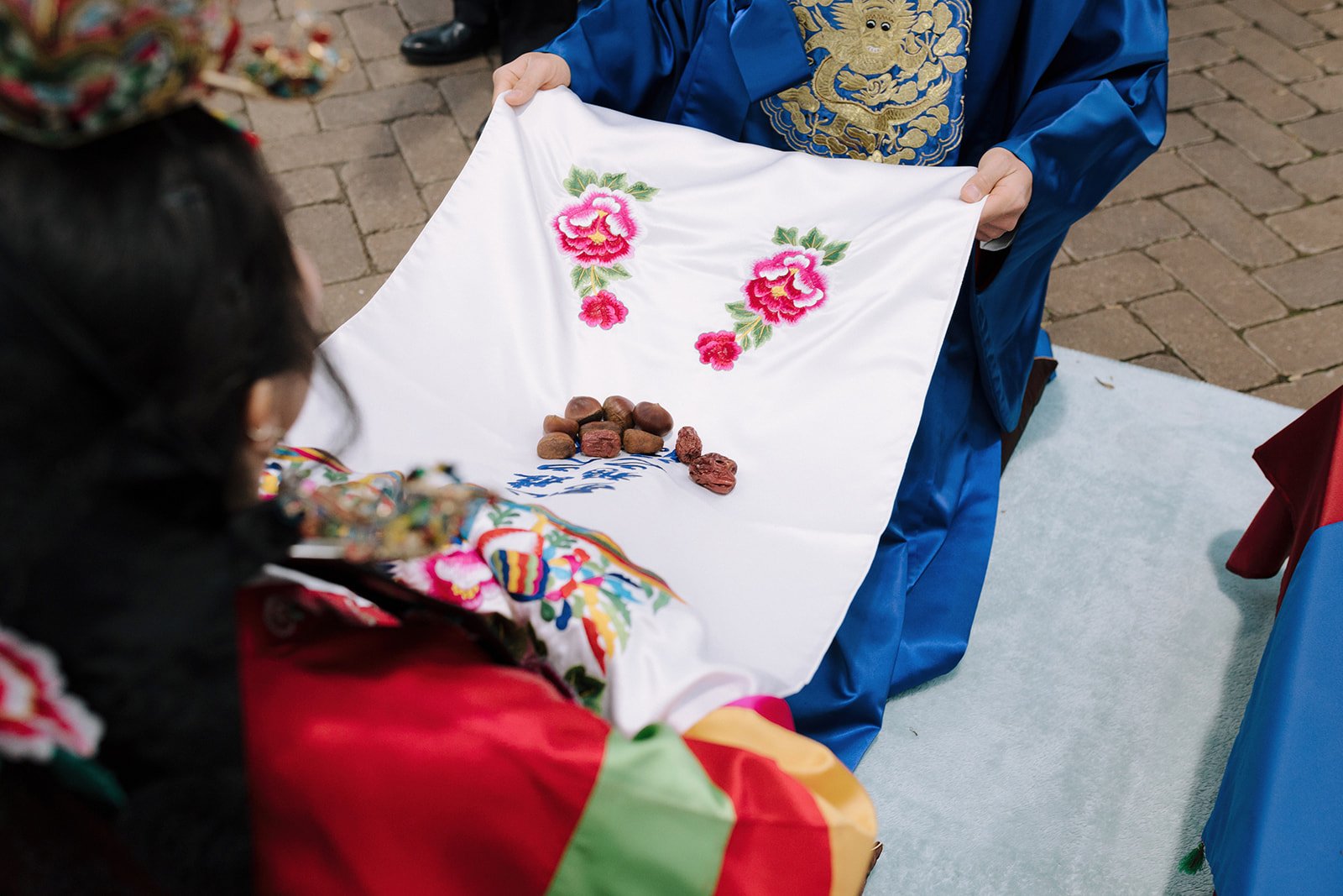 A person in traditional clothing holding a white embroidered cloth with pink floral patterns and stones on it, appearing to be part of a cultural or ceremonial event.