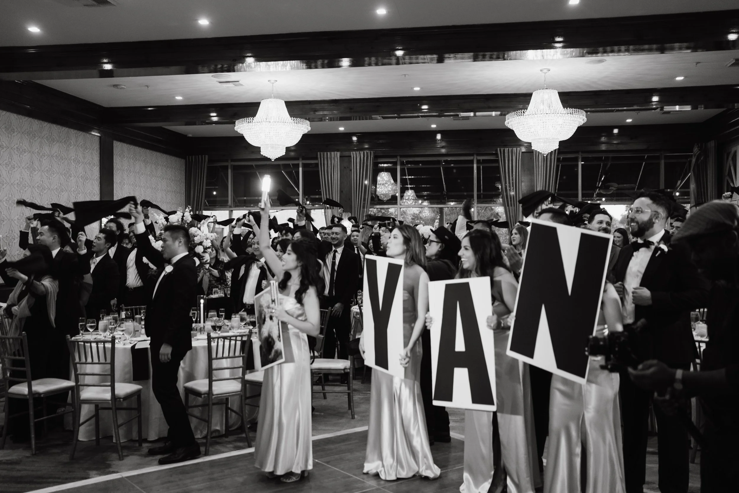 A large group of graduates in caps and gowns celebrating at a formal indoor graduation ceremony, some holding signs and raising their hands, with chandeliers hanging from the ceiling and decorated tables in the background.