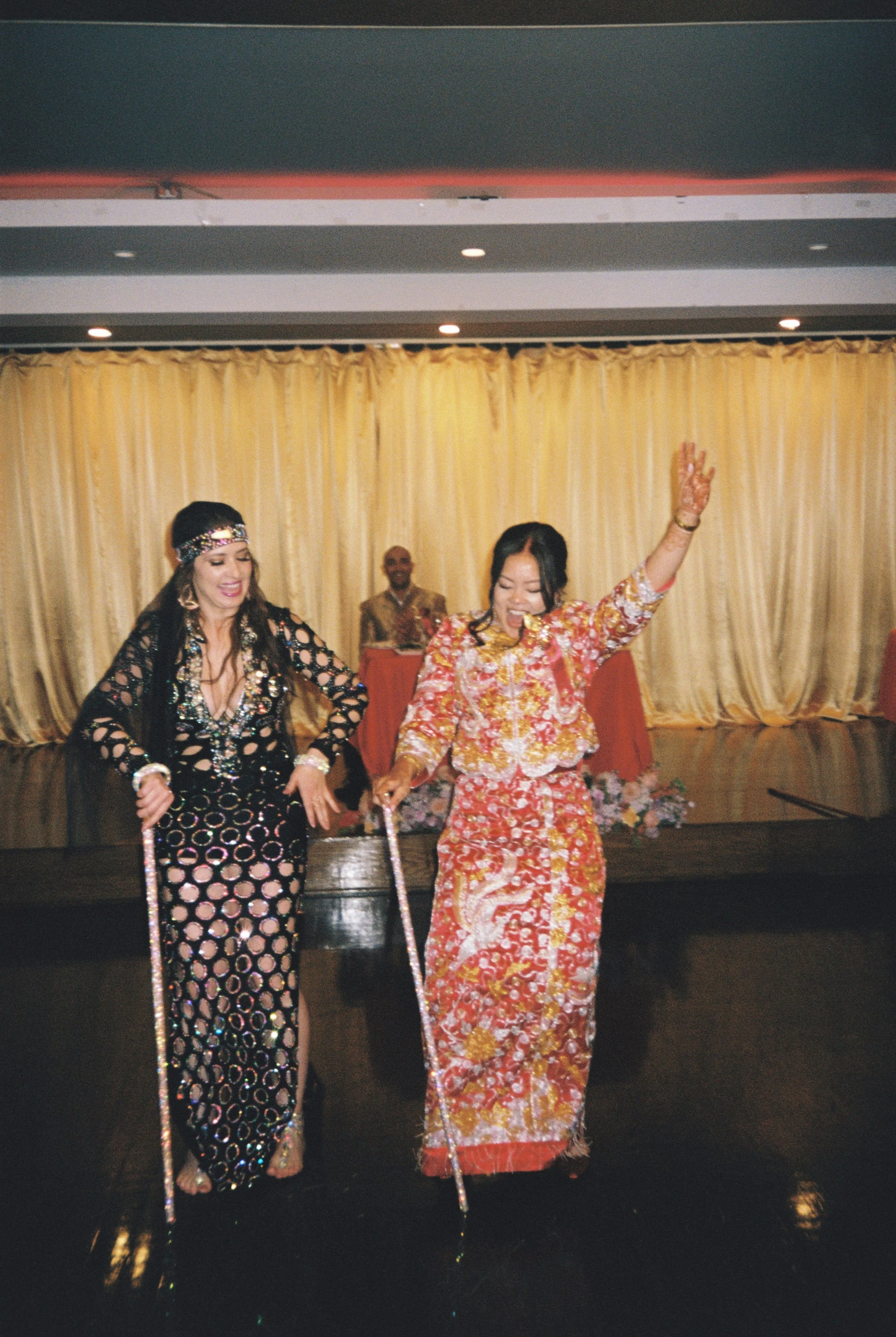 Two women dressed in colorful, one with a black dress with a pink and gold circular pattern and the other in a red floral dress, dancing with sticks on a stage with a cream curtain backdrop, accompanied by a man in the background.
