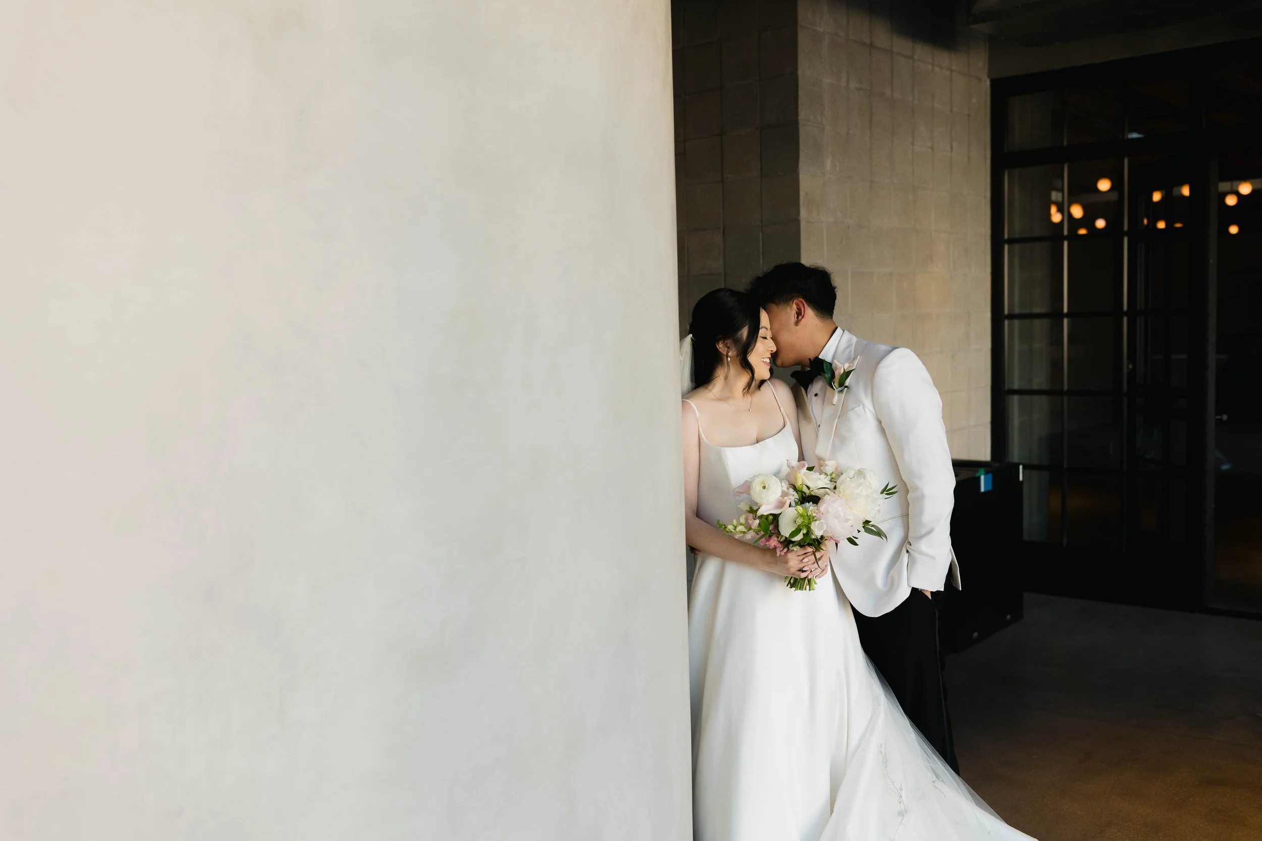 A bride and groom sharing an intimate moment, smiling with foreheads touching, standing close together with a bride holding a bouquet of white and pink flowers, in a modern indoor setting with large windows and warm lighting.