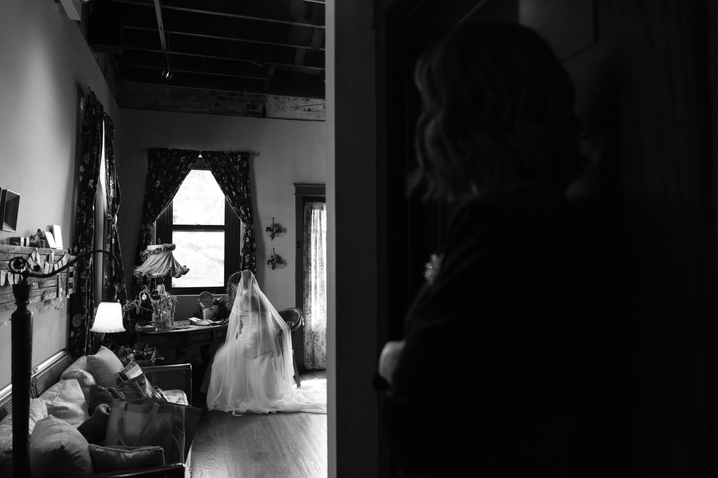 A black and white photo of a living room with a large window, curtains, a couch, and a table. There is a person in a wedding dress sitting on the floor with their back to the camera, looking at a small mirror. The image captures a cozy, rustic interi