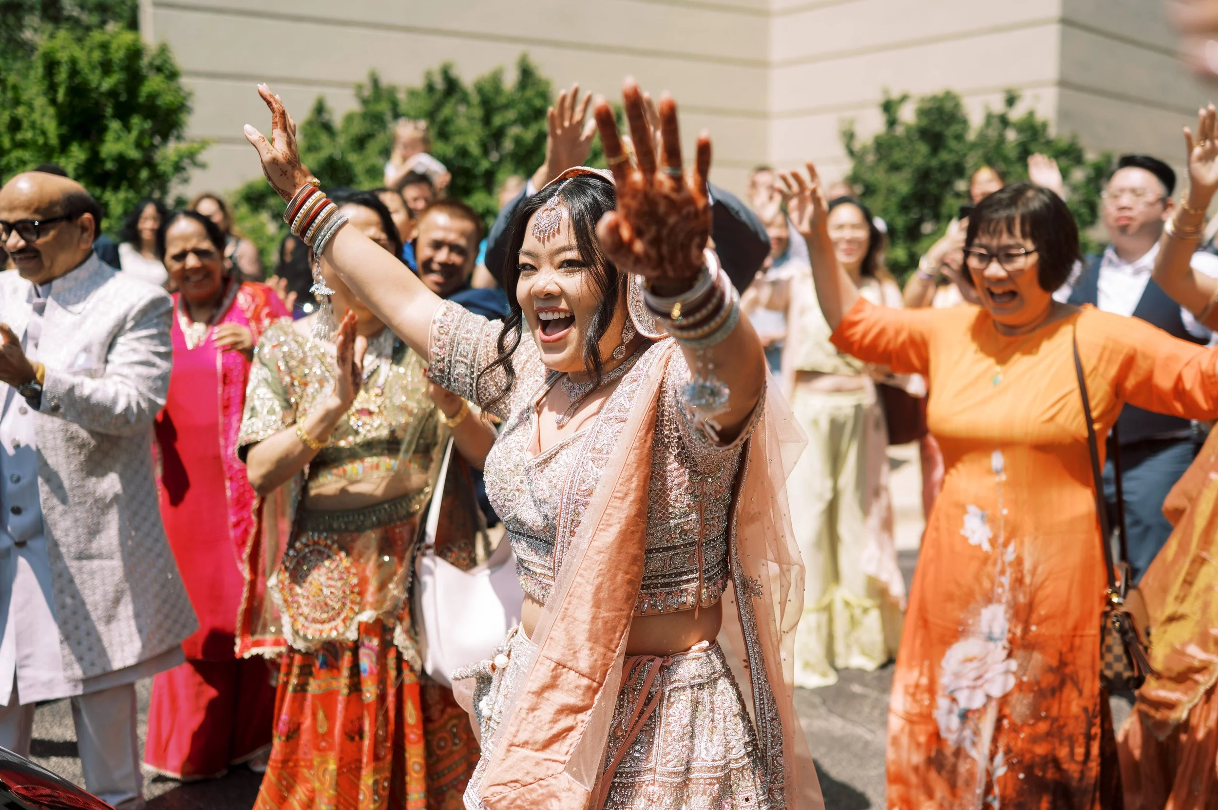 A vibrant outdoor celebration with diverse people dressed in traditional Indian attire, smiling and dancing in colorful clothes, surrounded by greenery.
