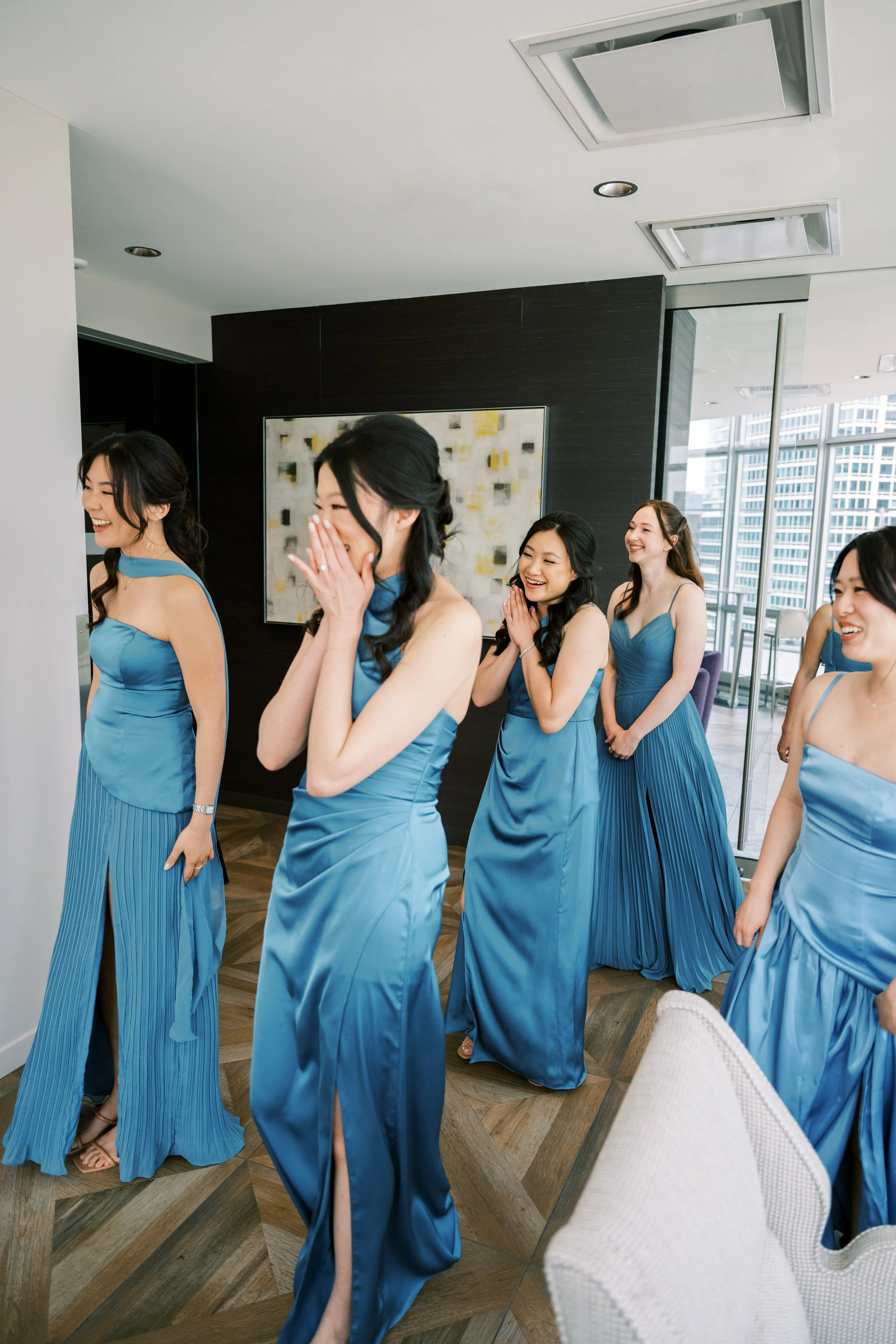A group of women in matching blue dresses with high slits, holding hands and smiling in a modern, high-rise apartment with large windows and a city view.