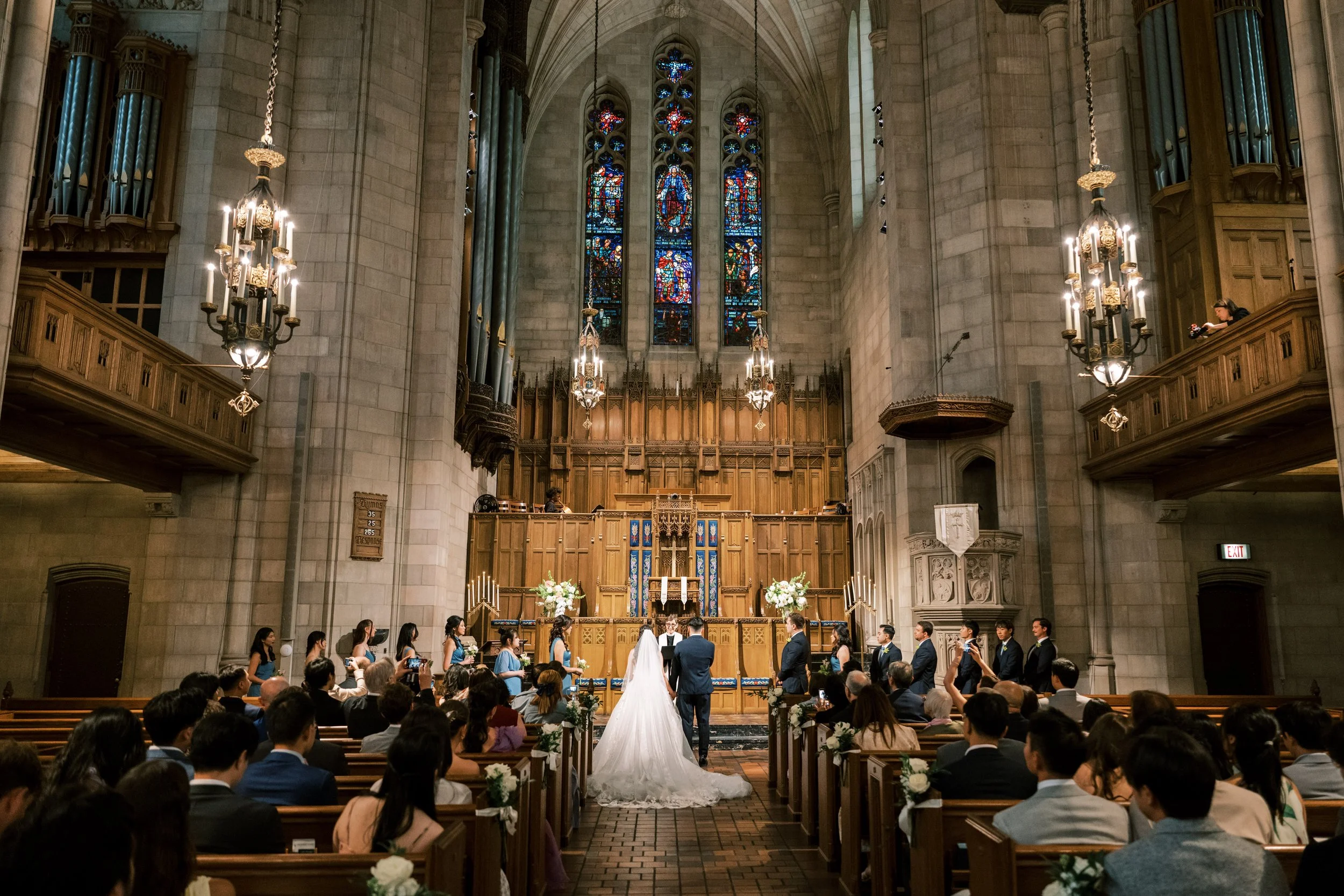 A group of people, including a bride in a white wedding gown and a groom in a tuxedo, standing at the altar in a grand church decorated for a wedding. They are surrounded by family and friends seated in pews, with beautiful stained glass windows and 