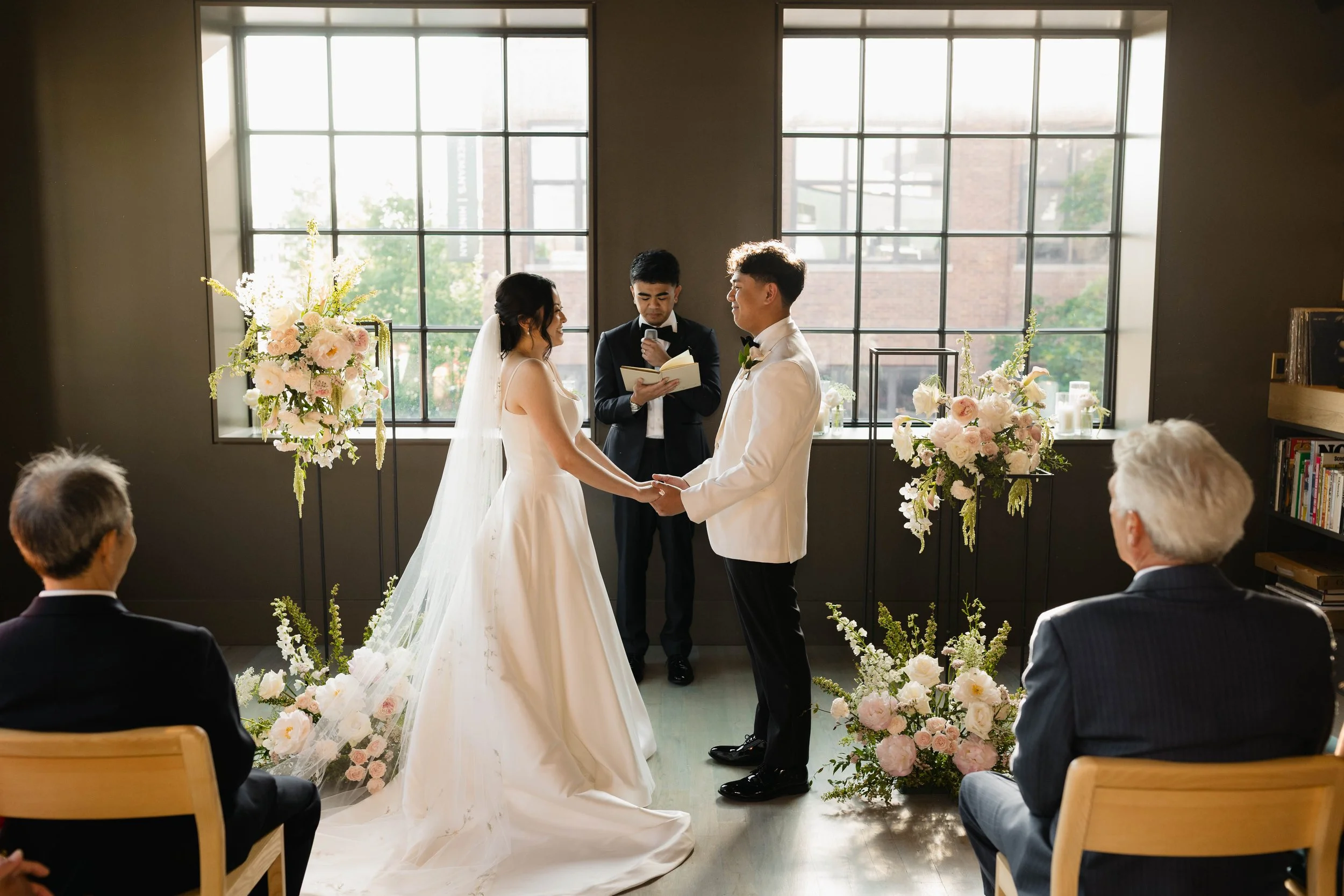 A wedding ceremony taking place indoors with two people holding hands and exchanging vows. The bride is wearing a white gown and veil, and the groom is dressed in a white tuxedo jacket with black pants. There is an officiant standing behind them read
