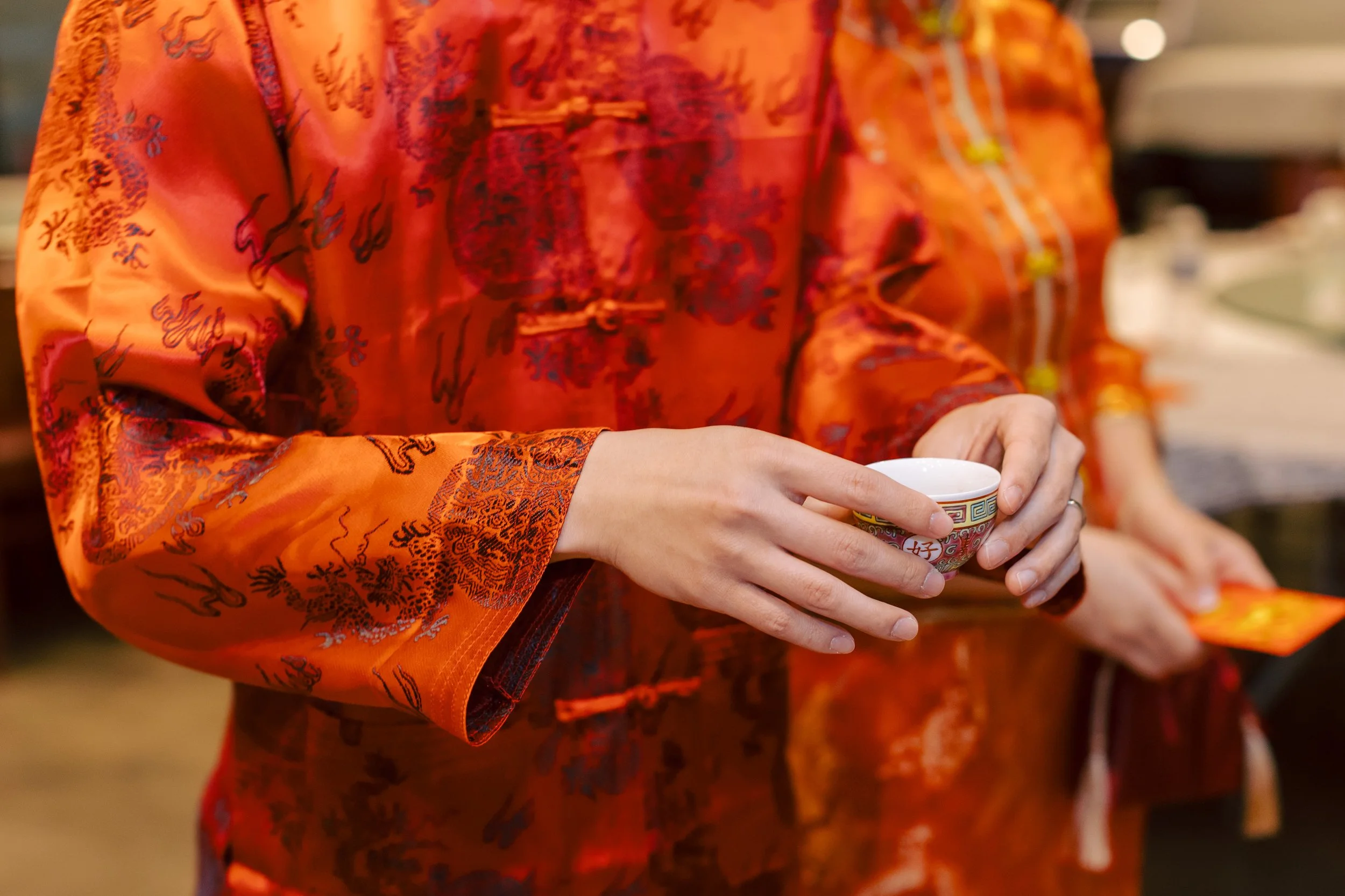 Person holding a small cup in a traditional Asian setting, wearing a bright orange robe with intricate patterns.