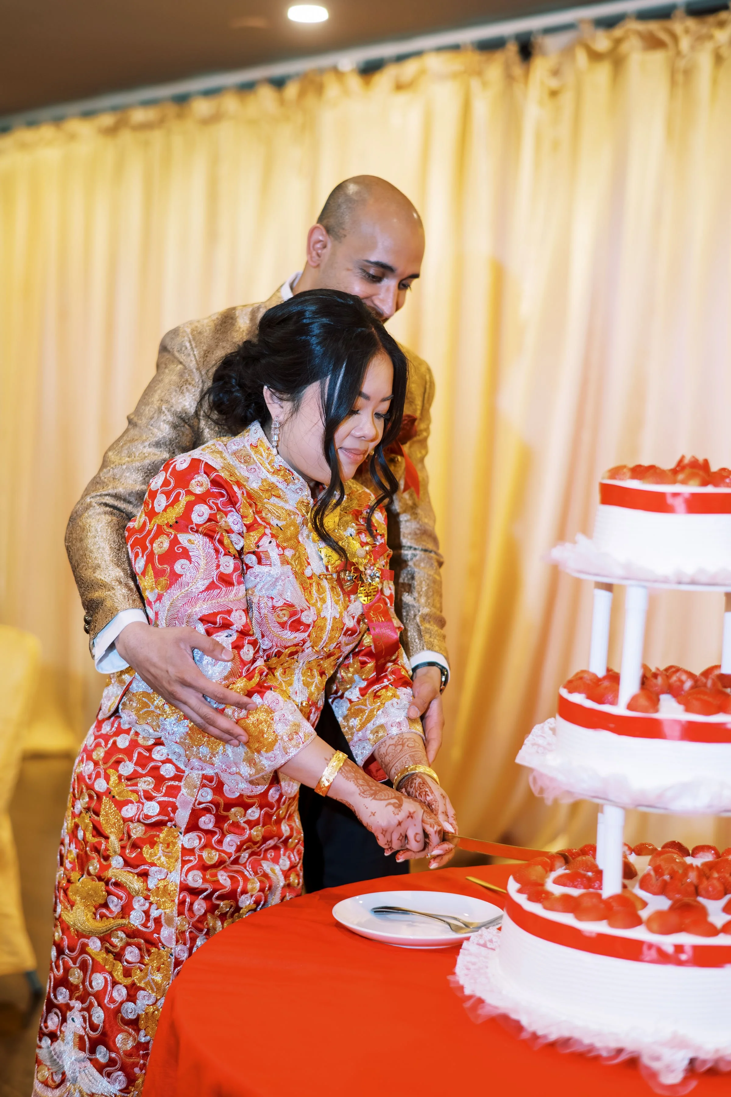 A man and woman in traditional Chinese wedding attire cutting a wedding cake at a celebration. The woman wears a red and gold dress with intricate patterns, and the man wore a gold-colored suit. There are yellow curtains in the background, and a tabl