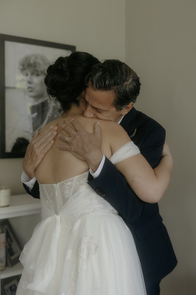 A man and a woman hugging tenderly, the woman in a wedding dress and the man in a dark suit, inside a room with framed black-and-white photo of a woman on the wall.