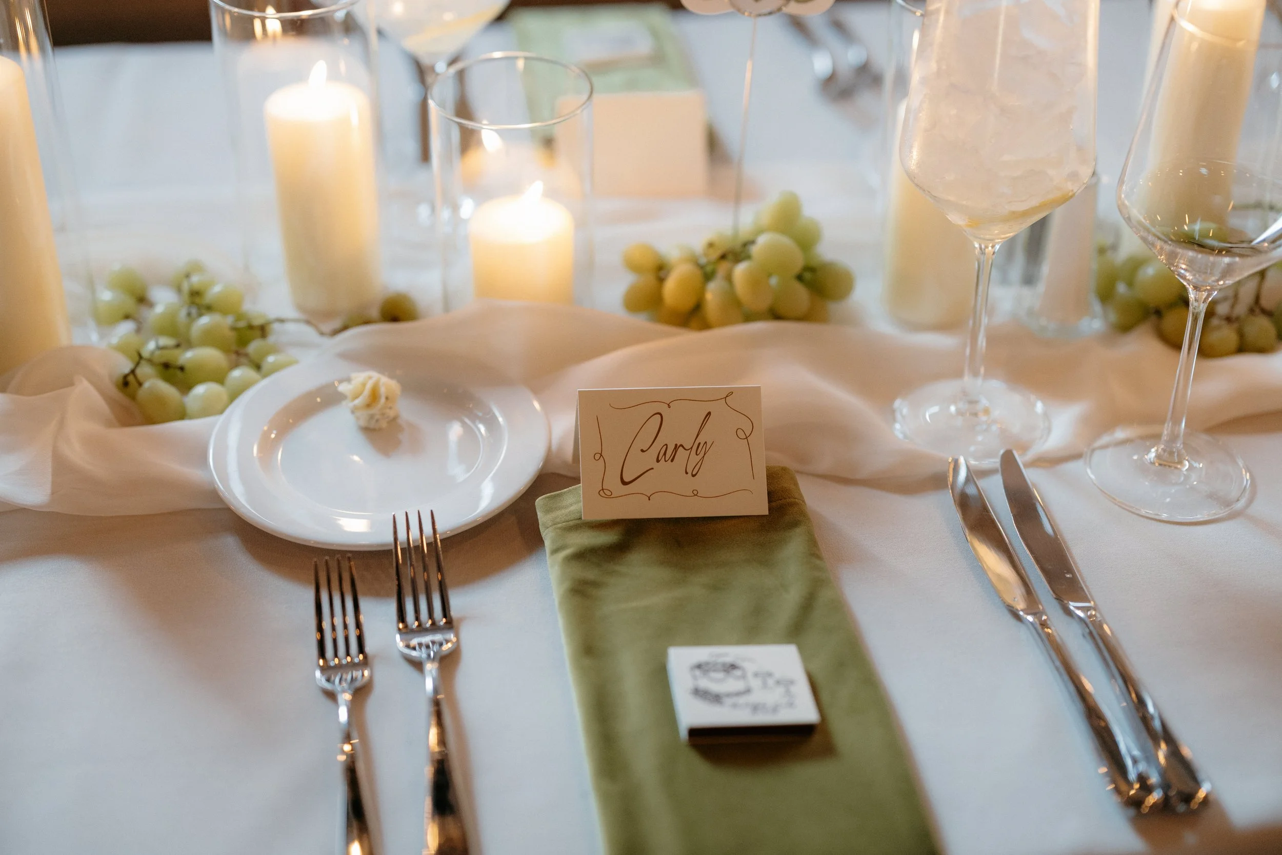 A decorated table with white candles, green grapes, and champagne glasses, with a place card that says 'Carly', set for a celebration or wedding reception.