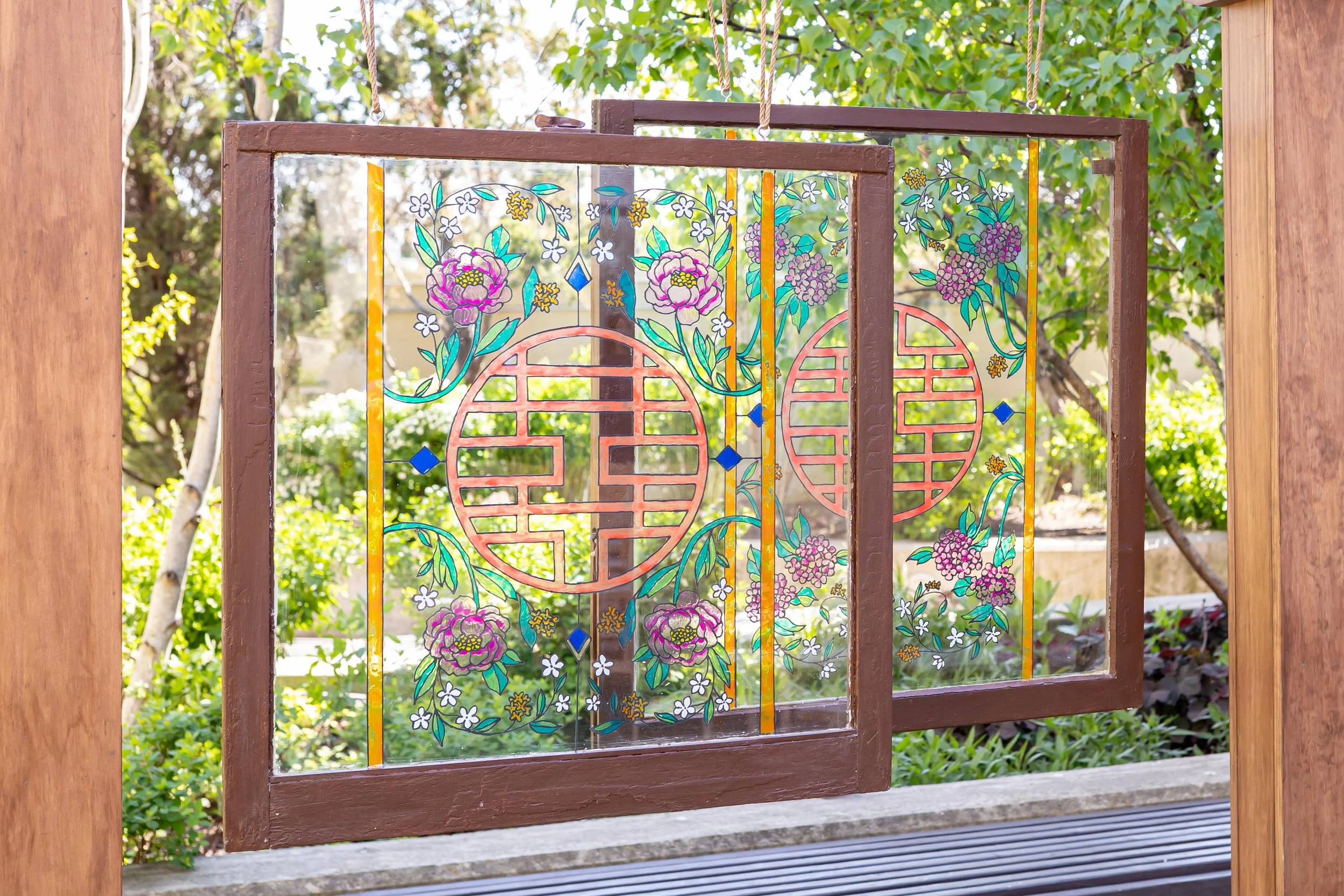 Decorative stained glass panel with floral design and circular red pattern, hanging on a wooden frame outdoors.