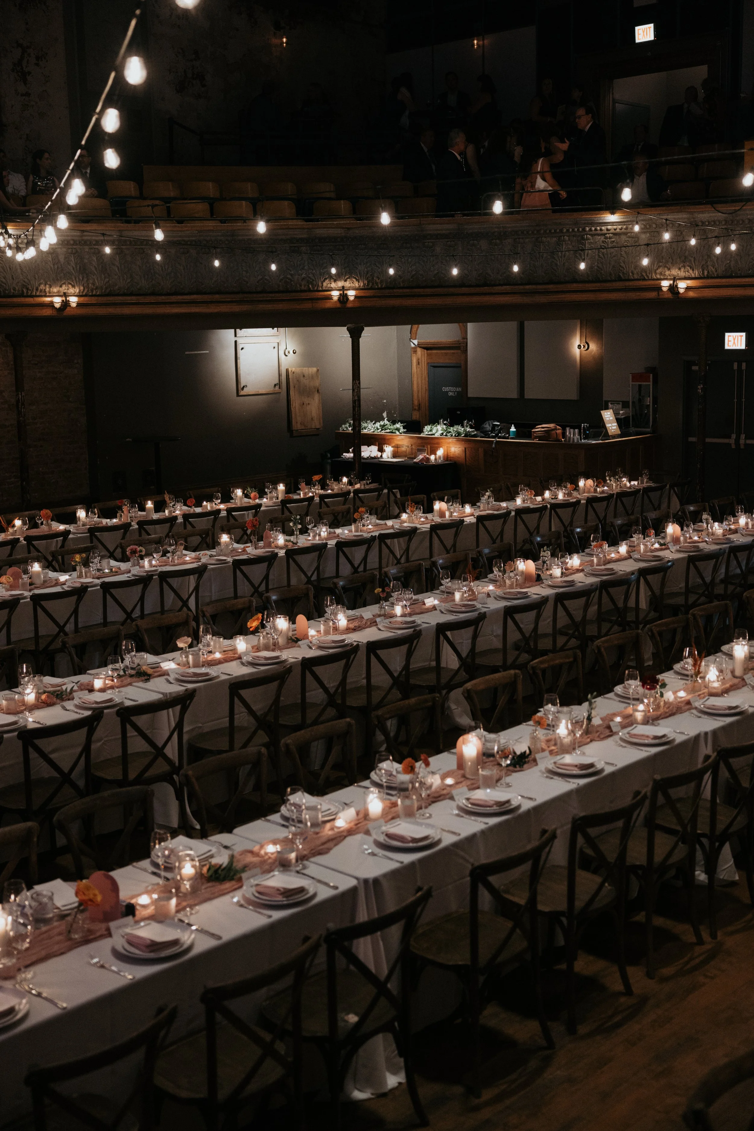 Banquet hall decorated with string lights, set with multiple tables arranged for a formal event, with chairs, candles, and floral centerpieces.