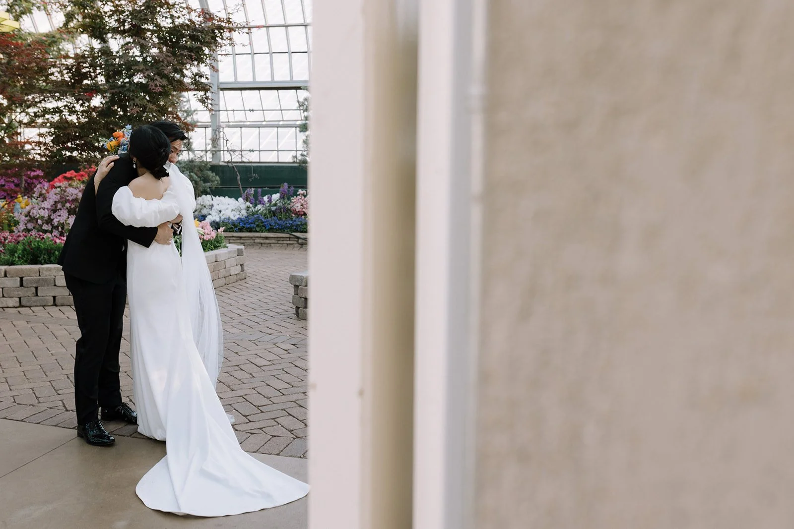 A bride and groom embrace each other inside a conservatory or tropical garden, surrounded by flowers and greenery, with a glass ceiling and brick pathway.