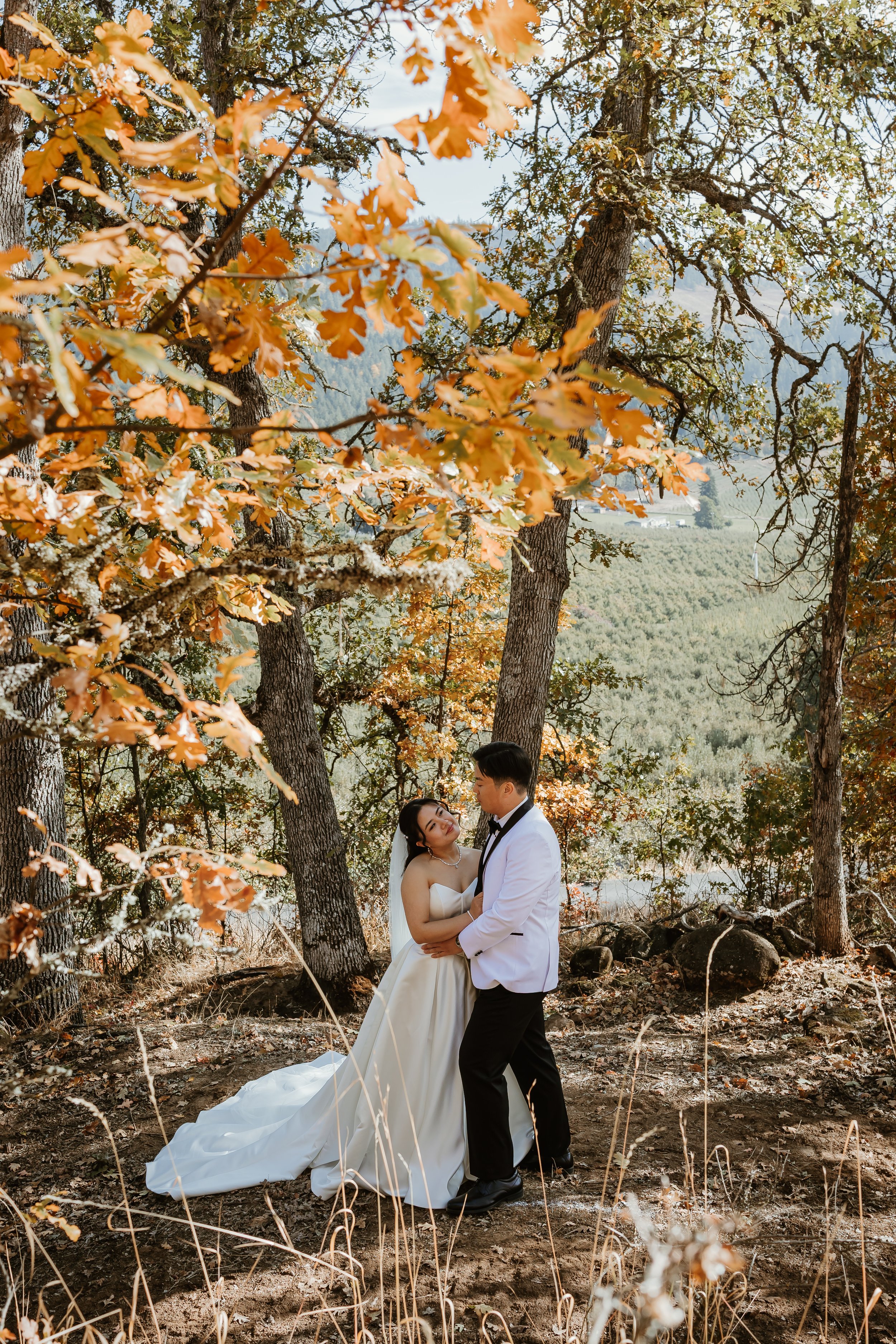A bride and groom are standing in a wooded area with fall foliage, holding each other and looking into each other's eyes, dressed in wedding attire.