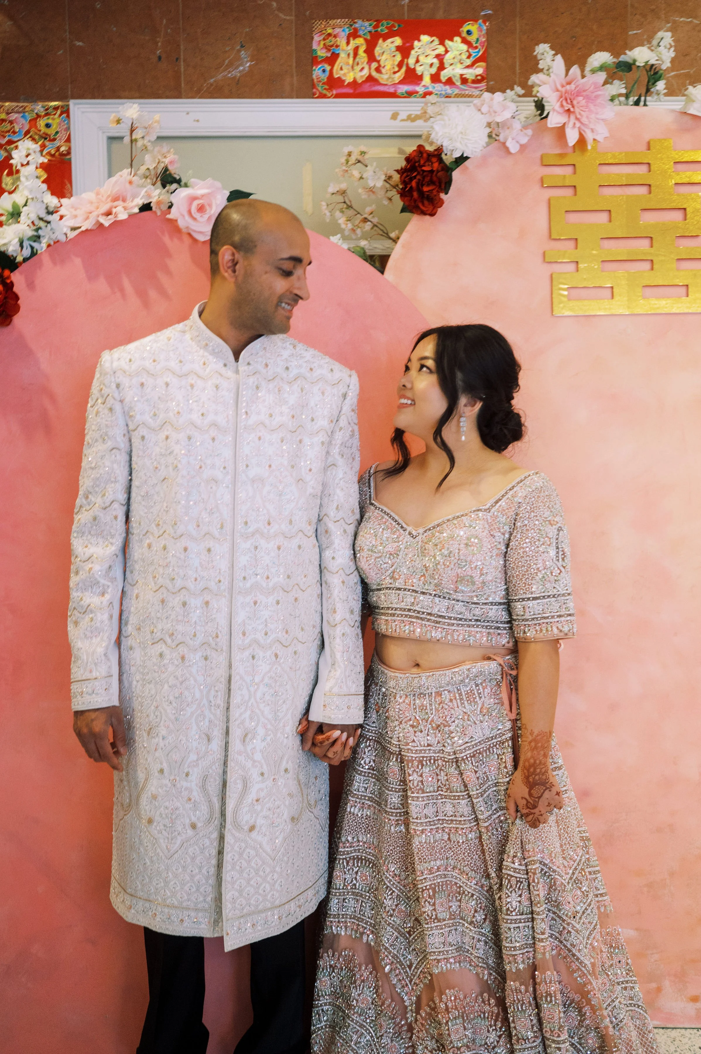 A couple dressed in traditional South Asian wedding attire holding hands, looking at each other and smiling, standing in front of a pink decorative backdrop with flowers and gold accents, celebrating a wedding.