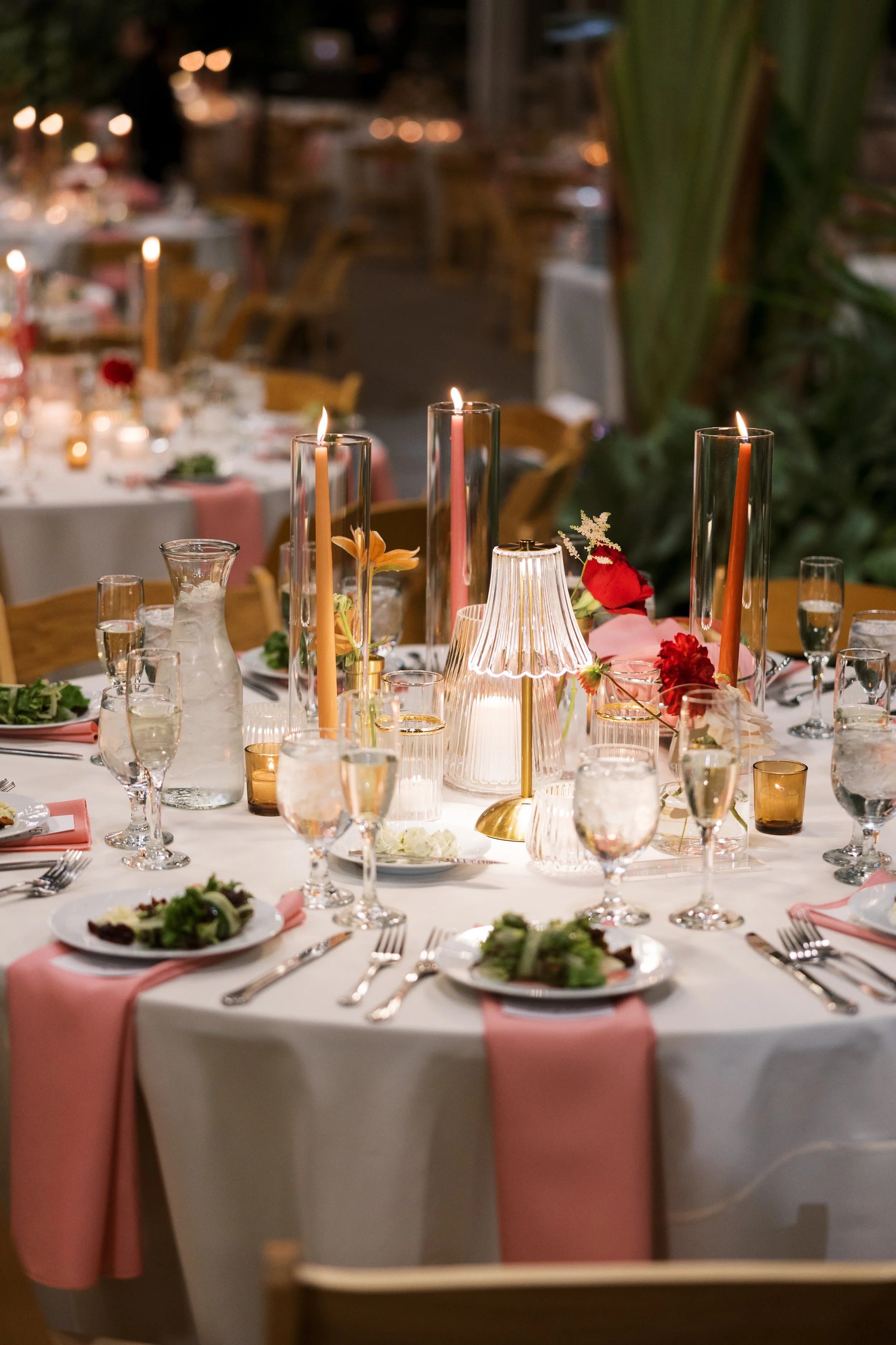 A decorated dining table set for a formal event with pink and white tablecloths, tall pink candles, floral arrangements, and tableware, with other tables and chairs visible in the background.
