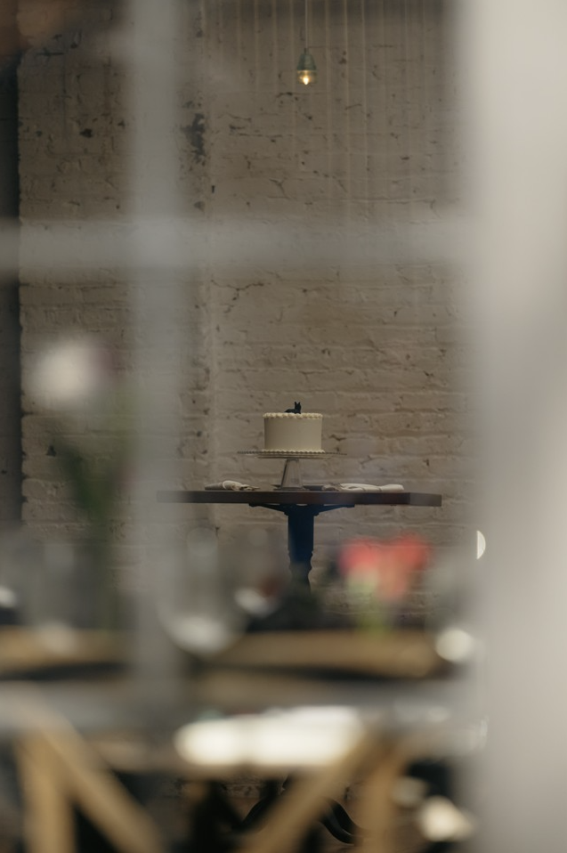 A small white cake with a black cat decoration on top placed on a table against a white brick wall, viewed through a blurred foreground.