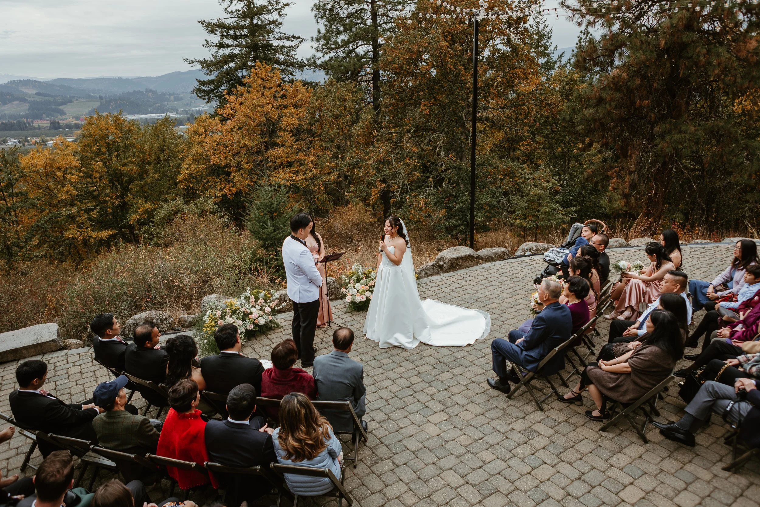 A wedding ceremony outdoors with trees showing fall foliage and distant mountains. A bride and groom stand before an officiant, surrounded by seated guests. The scene is set on a stone-paved area with flowers and rocks.