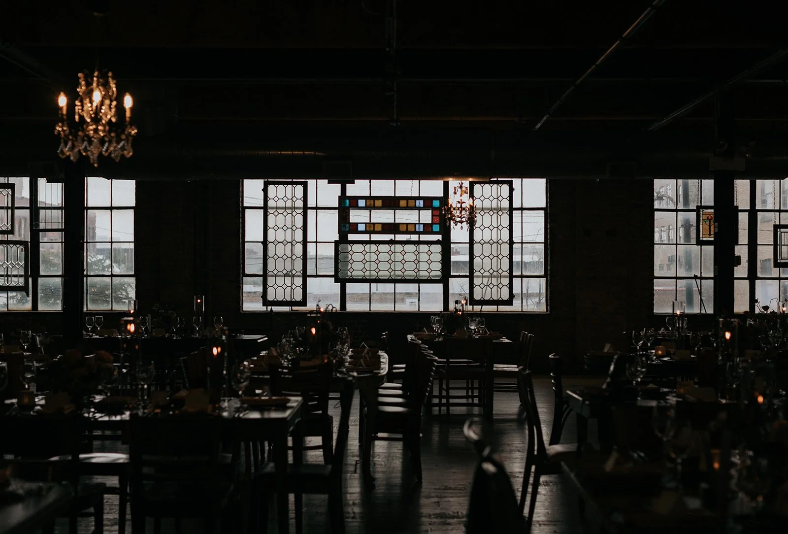 Dark interior of a restaurant with large windows, tables set with glassware, and chandeliers hanging from the ceiling.
