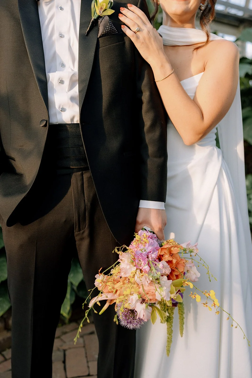 Close-up of a bride and groom on their wedding day, the bride touching the groom's shoulder, holding a colorful bouquet, with the groom dressed in a black tuxedo and the bride wearing a white wedding dress.