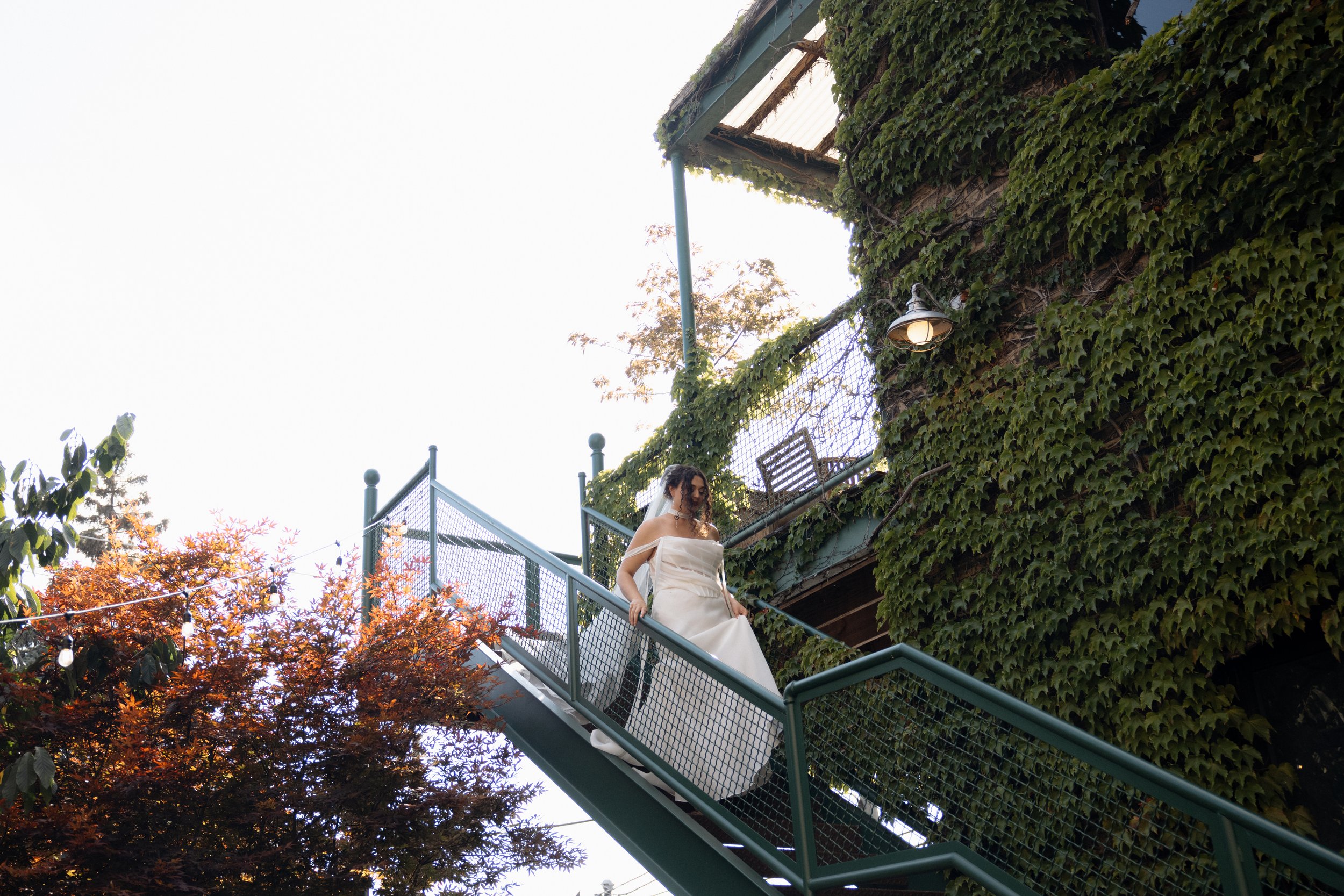 A bride in a strapless white wedding gown descending a green outdoor staircase with metal railings, surrounded by trees with autumn-colored leaves and a building covered in green ivy, outdoor street lamp, and a clear sky.
