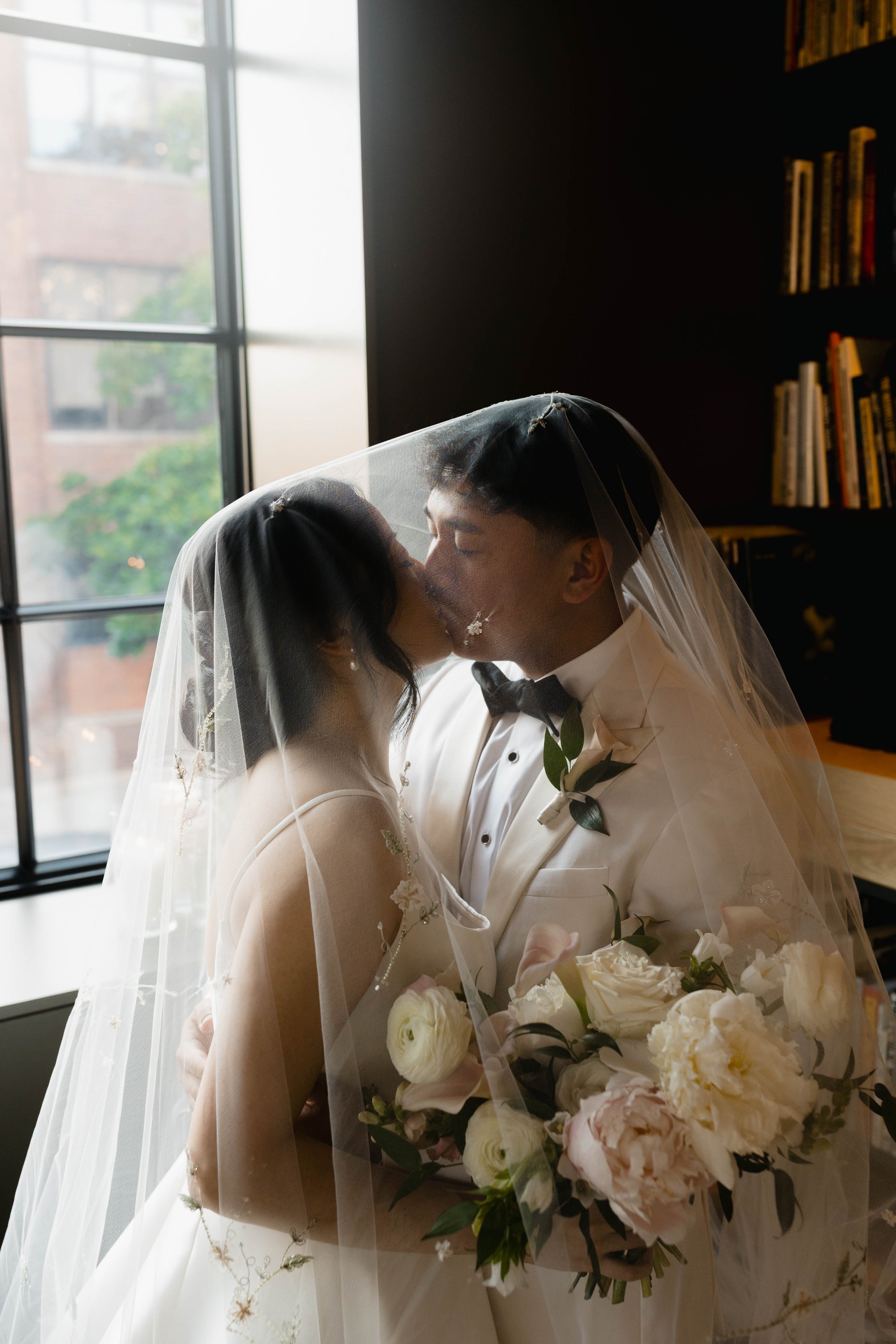 A bride and groom sharing a kiss indoors, with the bride holding a bouquet of white and pink flowers, and a window with natural light in the background.