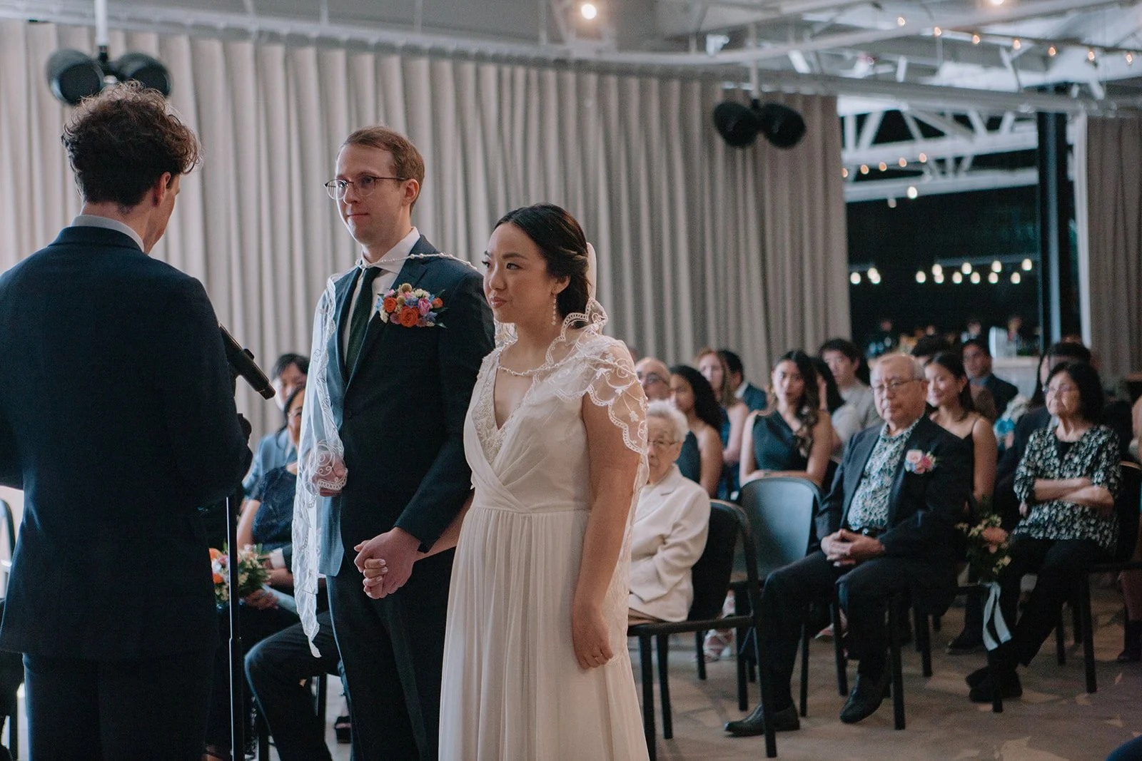 A wedding ceremony taking place indoors with a bride, groom, and officiant. The bride is wearing a white dress, and the groom is dressed in a dark suit with a boutonniere. Guests sit in the background watching the ceremony.