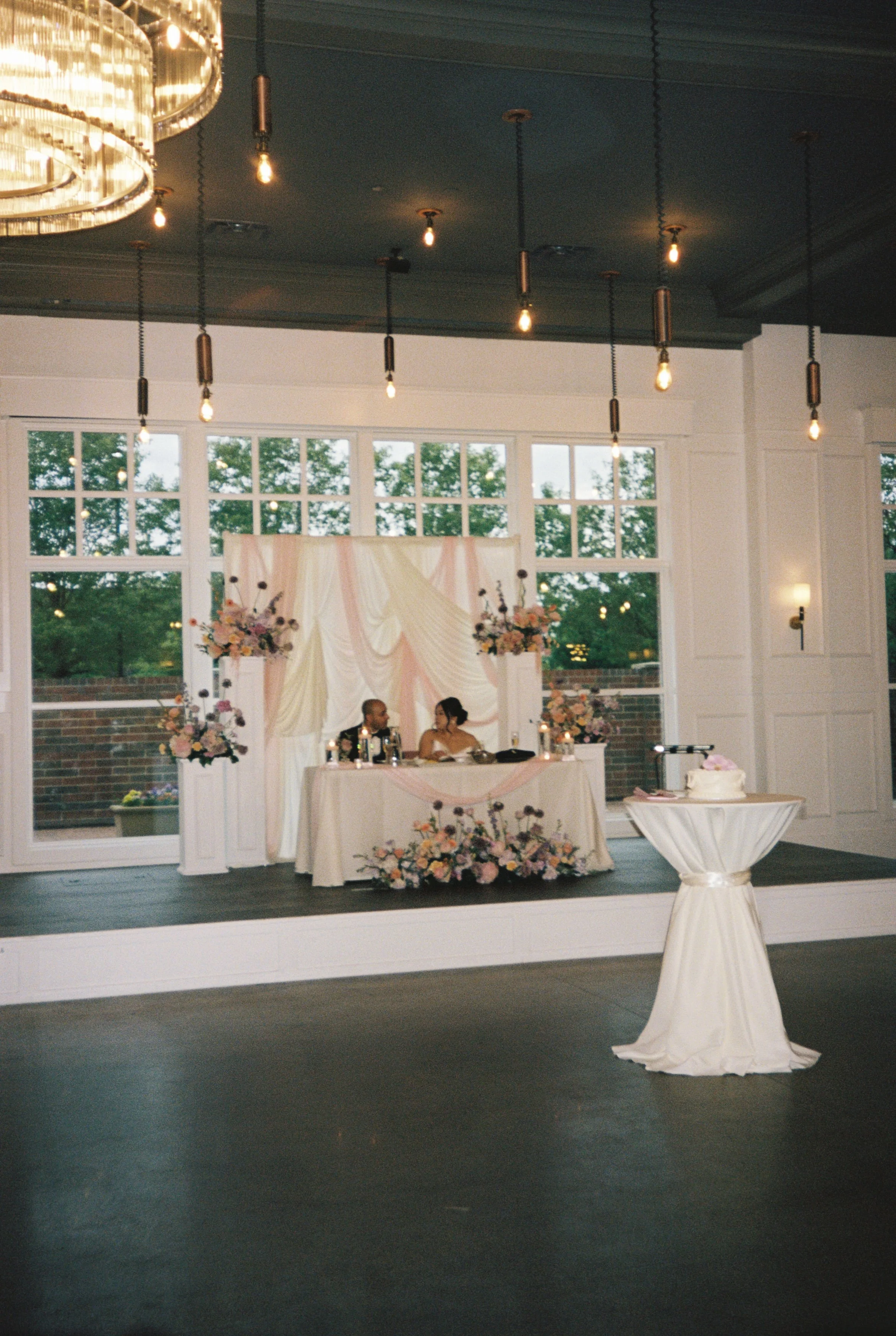 Wedding reception area with a decorated sweetheart table for the bride and groom and floral arrangements, set against large windows showing outdoor greenery, featuring hanging light bulbs and a small round table with a white cloth in the foreground.