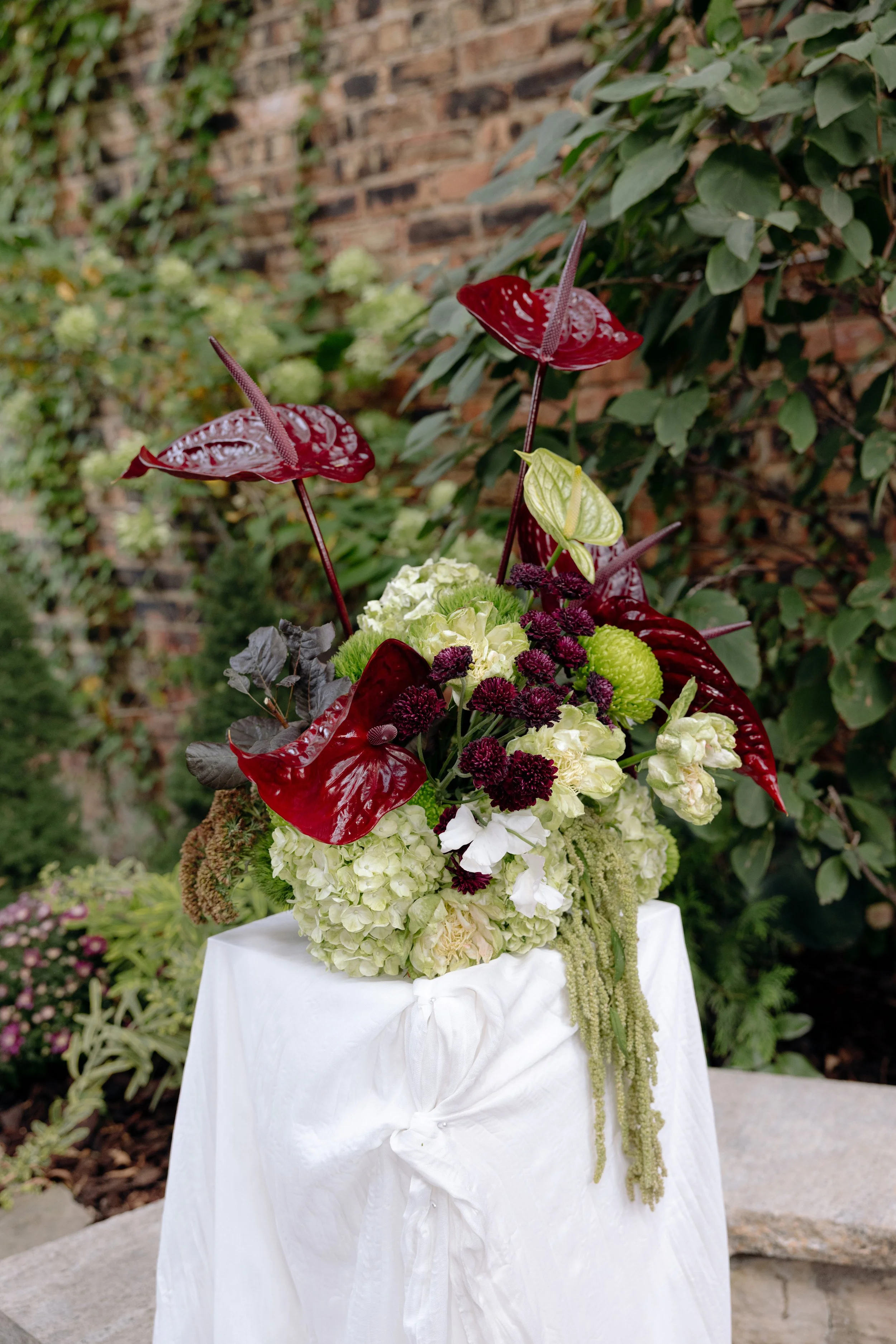 A floral arrangement featuring red anthuriums, white hydrangeas, and greenery, displayed on a white tablecloth outdoors with a brick wall and plants in the background.