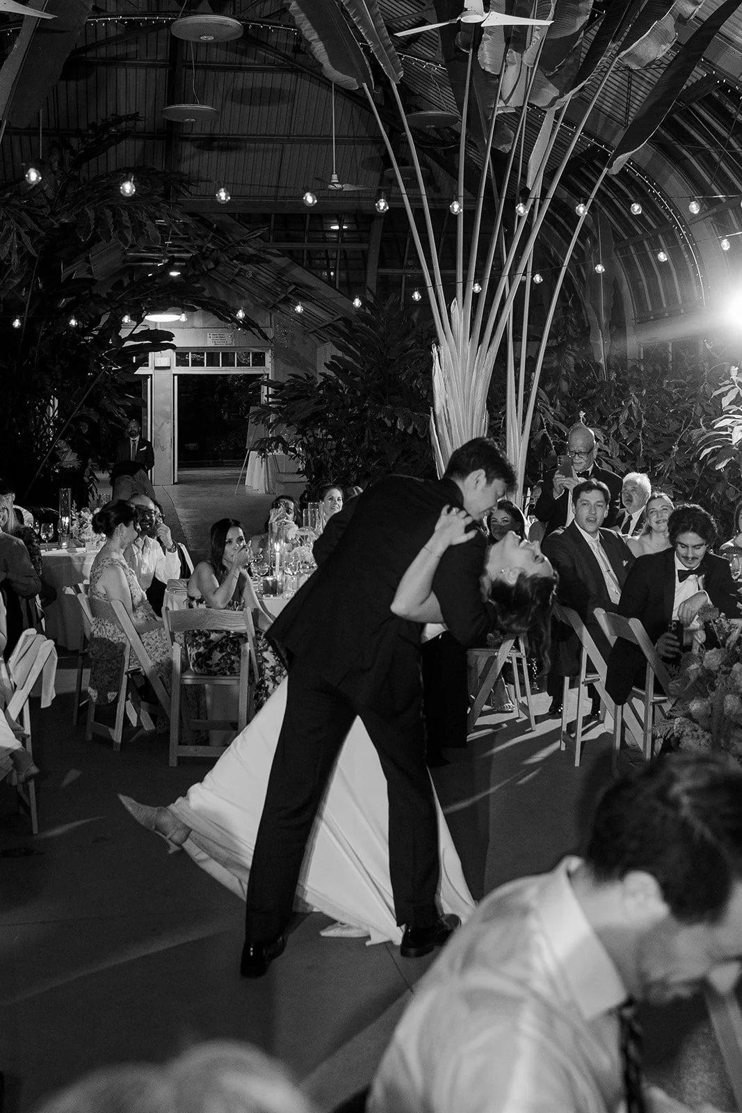 A couple dancing closely at a wedding reception, surrounded by seated guests in a decorated indoor venue with string lights and large plants.