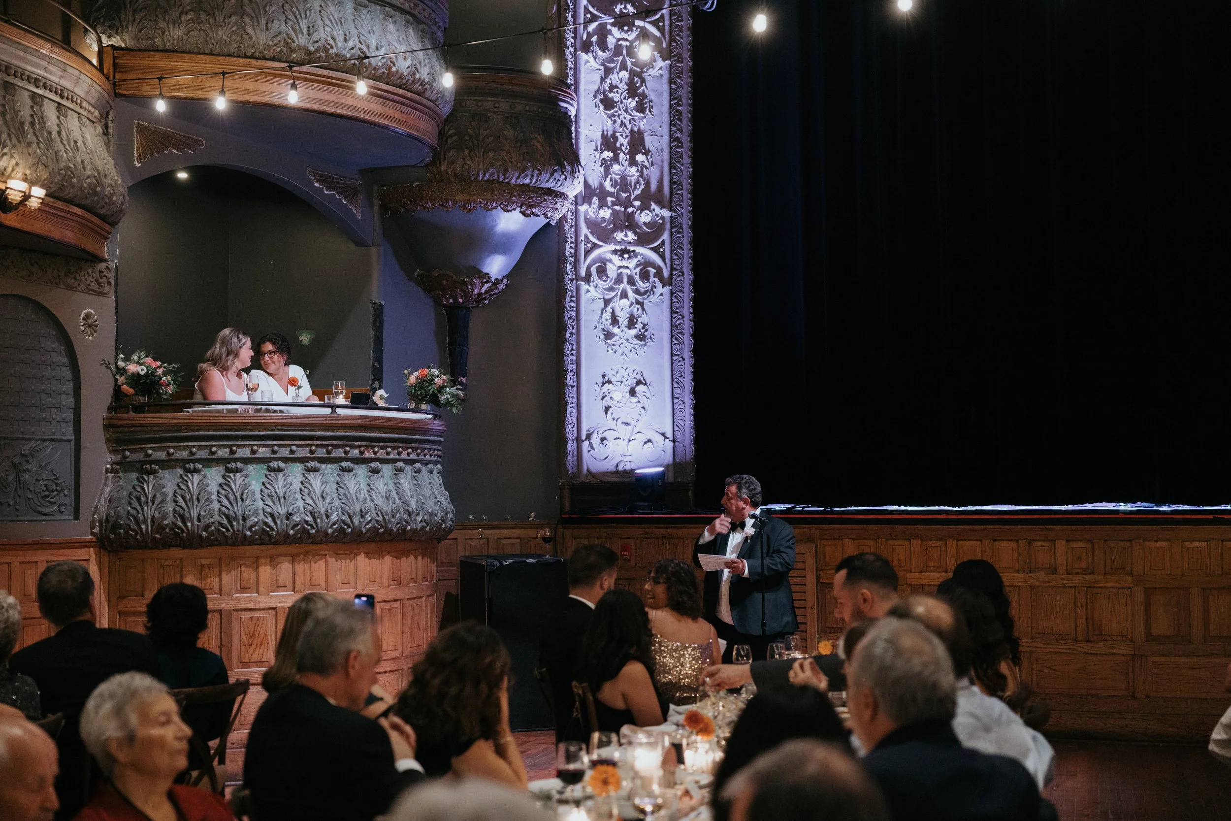 A formal dinner event in a theater with ornate architectural details. Two women are seated in a balcony box, talking, while a man in a tuxedo is standing below, reading from a paper. The audience is seated at tables with flowers and drinks, watching 