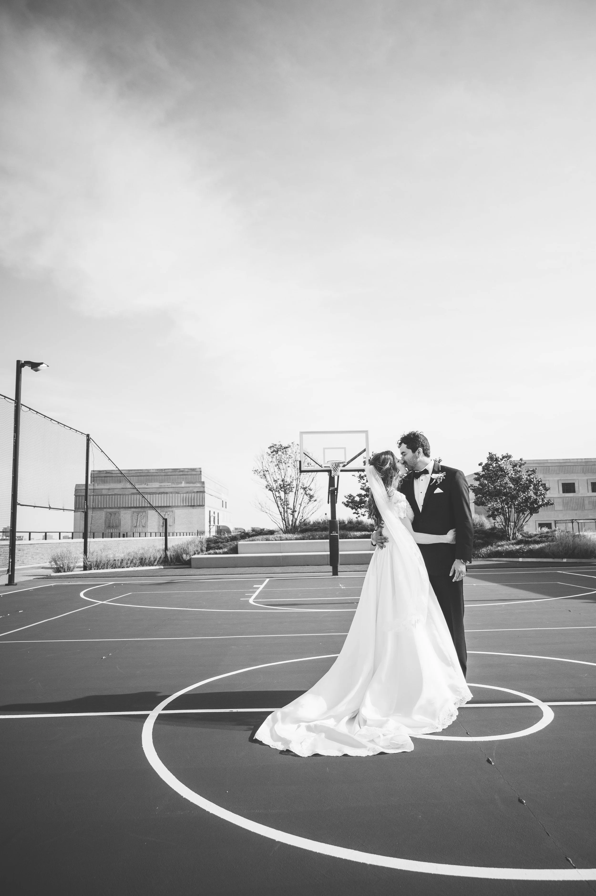 A bride and groom dressed in wedding attire are standing on a basketball court, sharing a kiss.