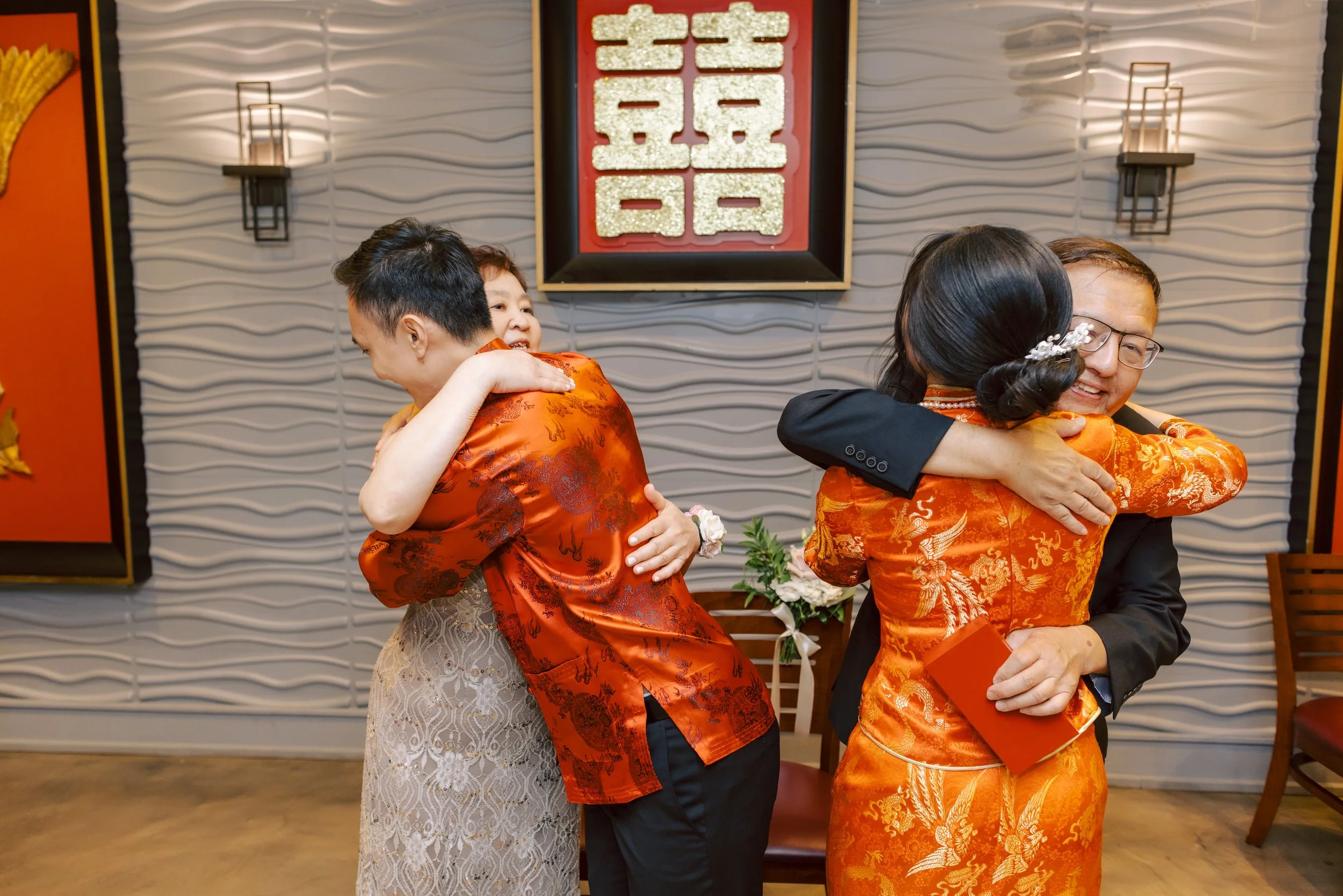 Four people are hugging each other at an event. Two women and two men, all dressed in traditional Asian attire, are embracing in pairs. The background features a decorative wall with Chinese characters and lantern-style wall sconces, indicating a cel