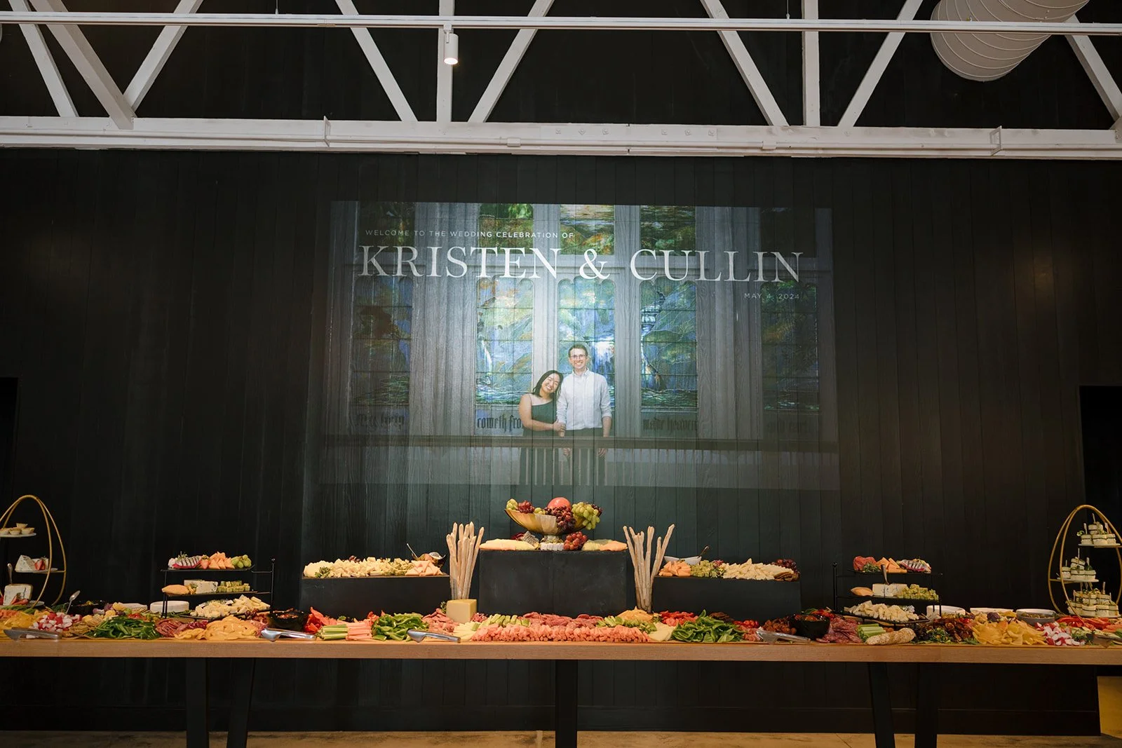 Wedding reception with a large buffet table of food, a black wall with a large wedding photo and text, and a high ceiling with white beams.