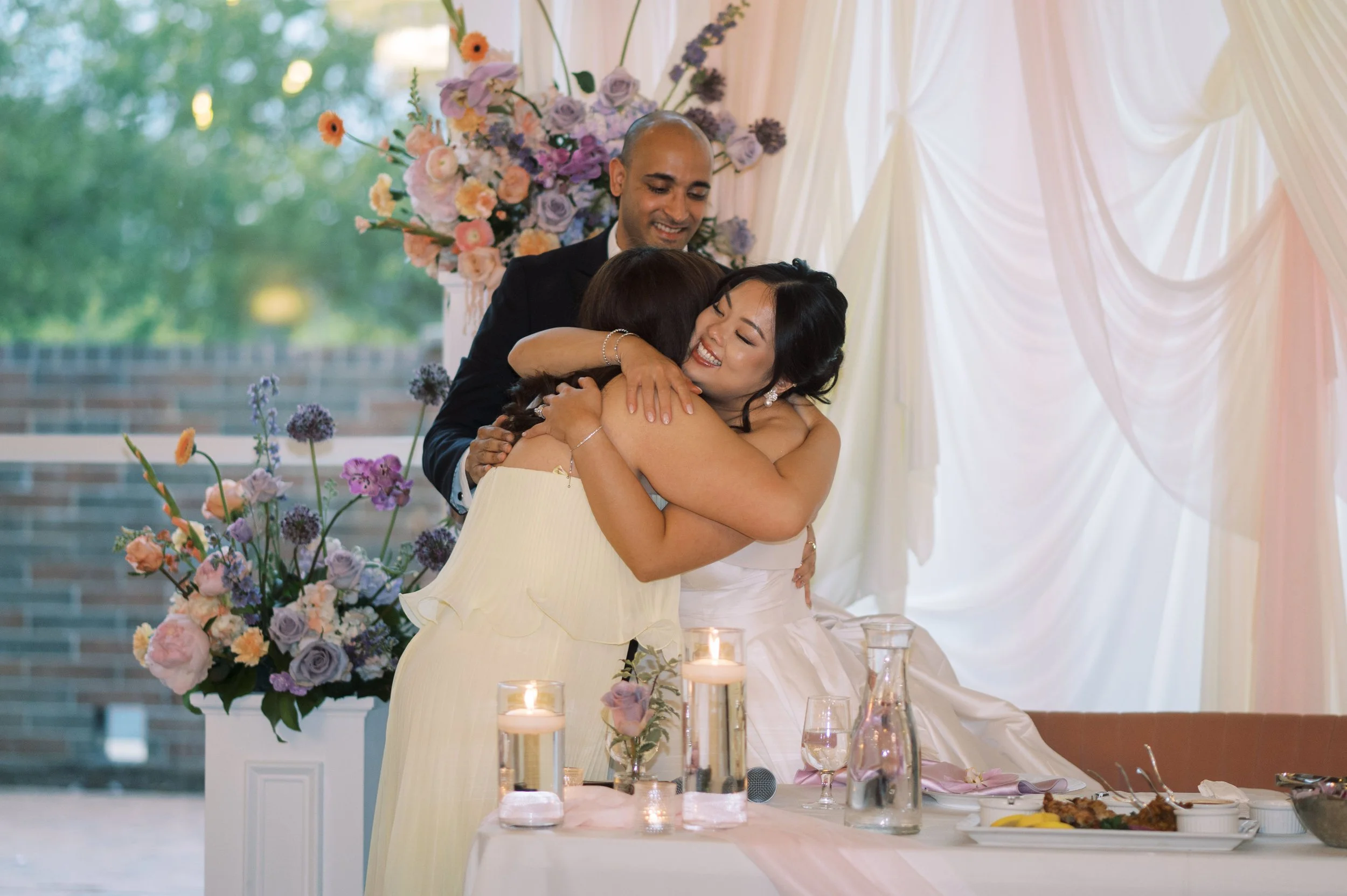 A man in a suit and a woman hugging at a wedding reception, with a floral arrangement and candles on the table, and decorative drapes in the background.