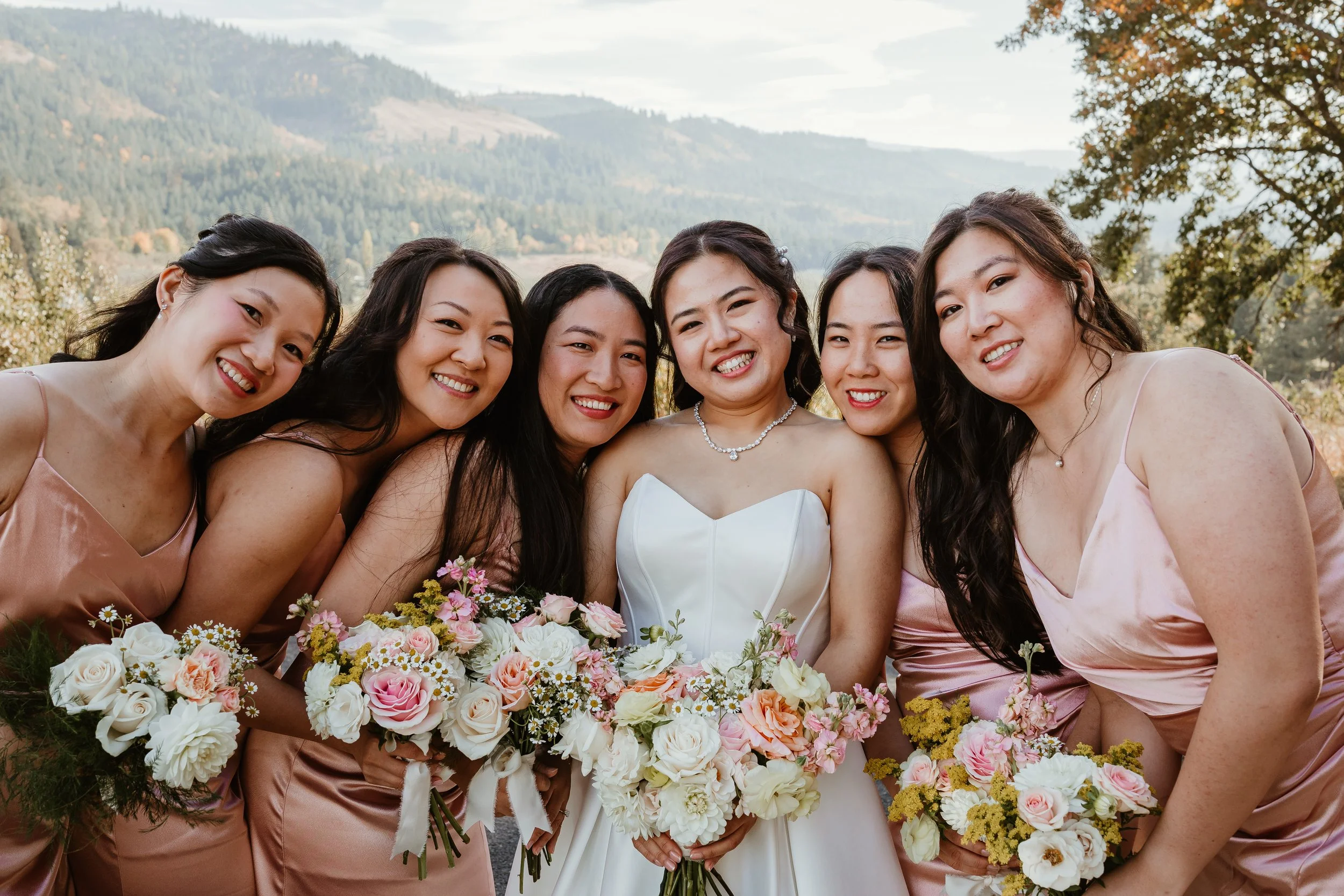 A group of six smiling women at a wedding, wearing pink dresses or a white wedding gown, holding bouquets of flowers, outdoors with trees and mountains in the background.