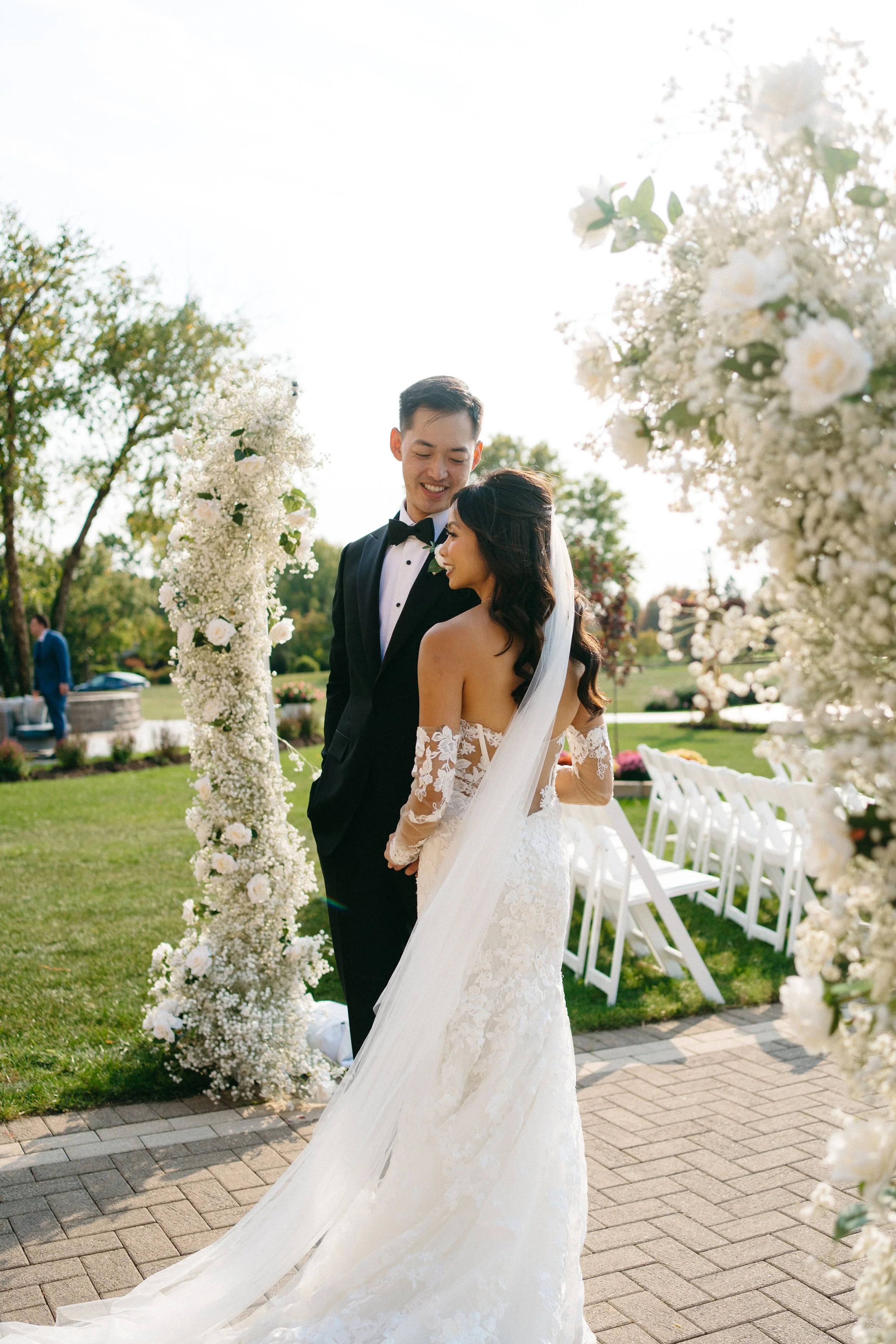 A bride and groom smiling and holding each other outdoors during a wedding ceremony, surrounded by floral decorations and white chairs.