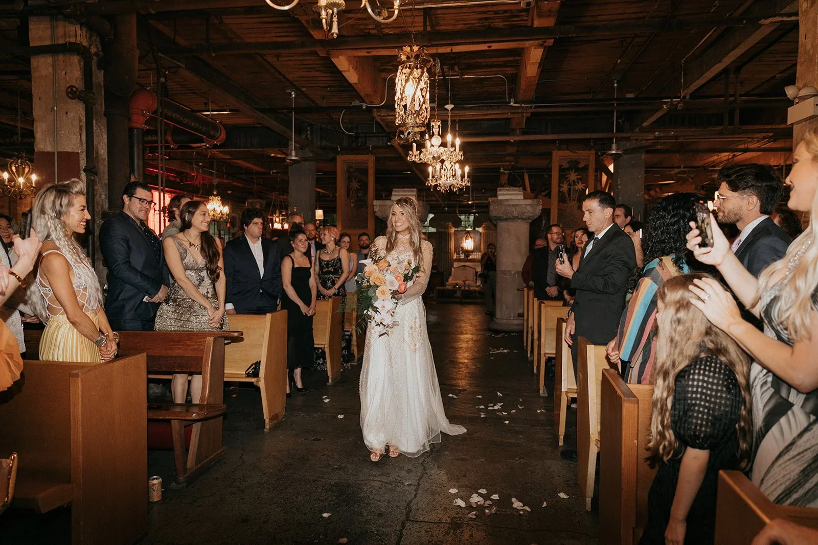 A bride walking down the aisle holding a bouquet of flowers in a wedding ceremony inside a rustic venue with chandeliers and wooden decor, surrounded by guests on both sides capturing the moment.