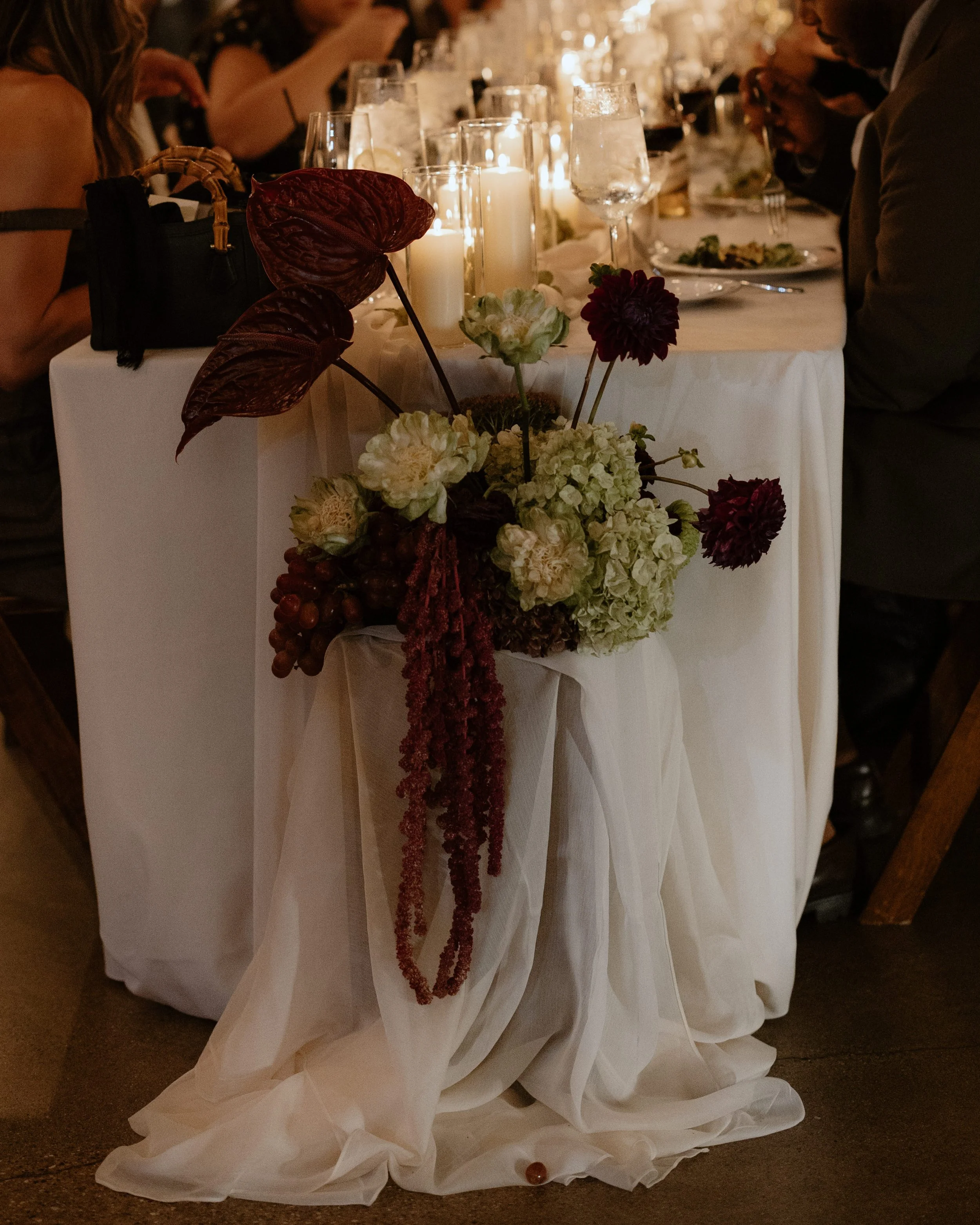 A wedding reception table setting with candles and floral arrangements. The table features plates, glasses, and silverware, and guests are seated around it. The focus is on an elegant floral centerpiece with white and dark red flowers, and a red leaf