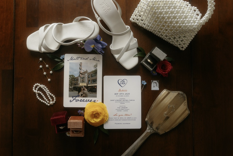 Flat lay of wedding accessories including white high-heeled sandals, a pearl handbag, pearl jewelry, a yellow flower, a wedding invitation, a photograph, small velvet rings boxes, a lipstick, a flower, and a mirror on a wooden surface.