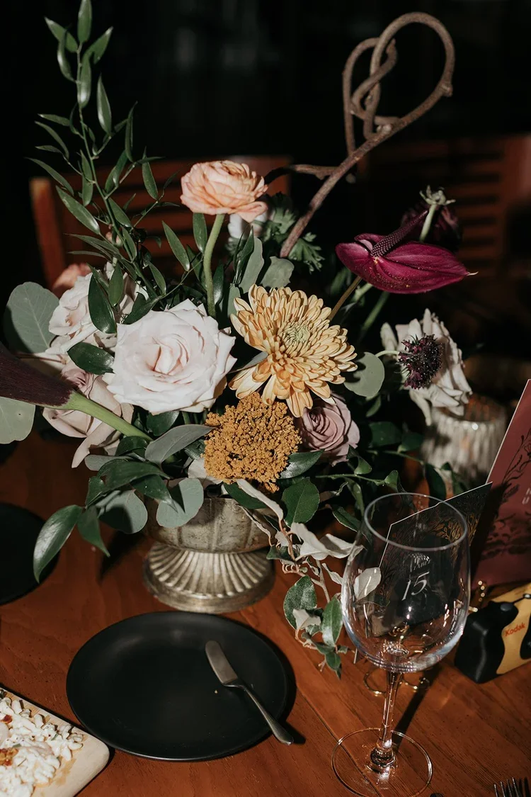 A floral centerpiece with various flowers and greenery on a wooden table, accompanied by a black plate with a spoon, an empty wine glass, and a decorated card, in a dimly lit setting.