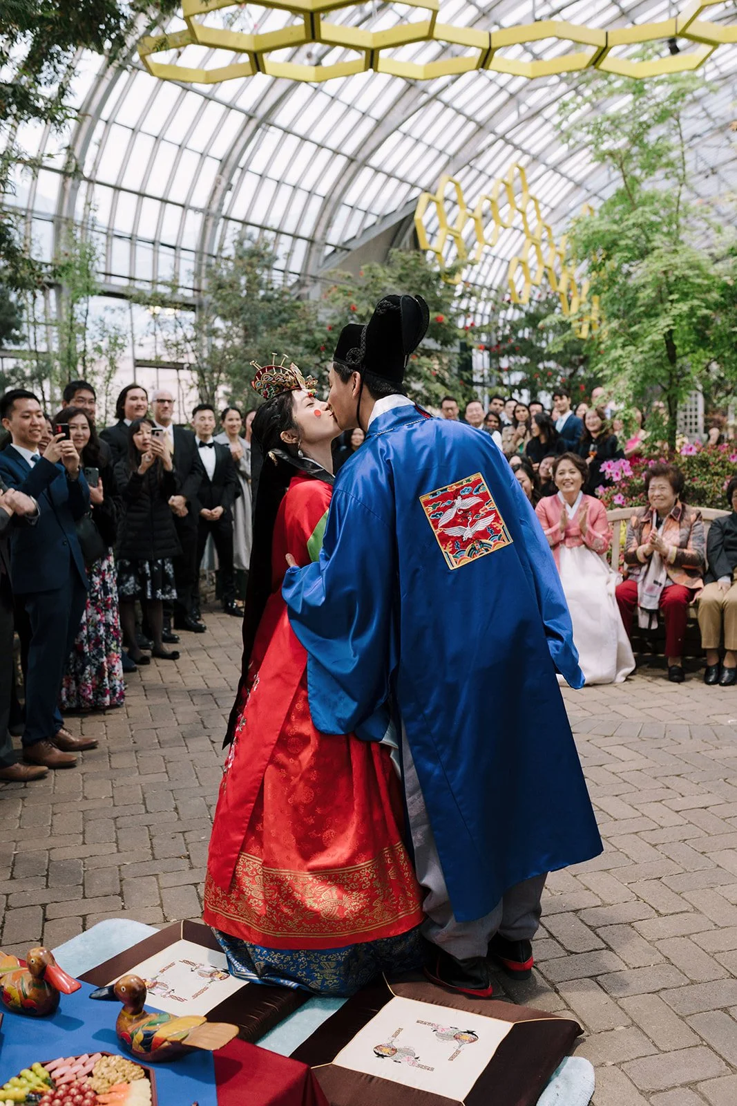A couple in traditional Korean clothing sharing a kiss during a wedding ceremony in a greenhouse, with onlookers taking photos and smiling.