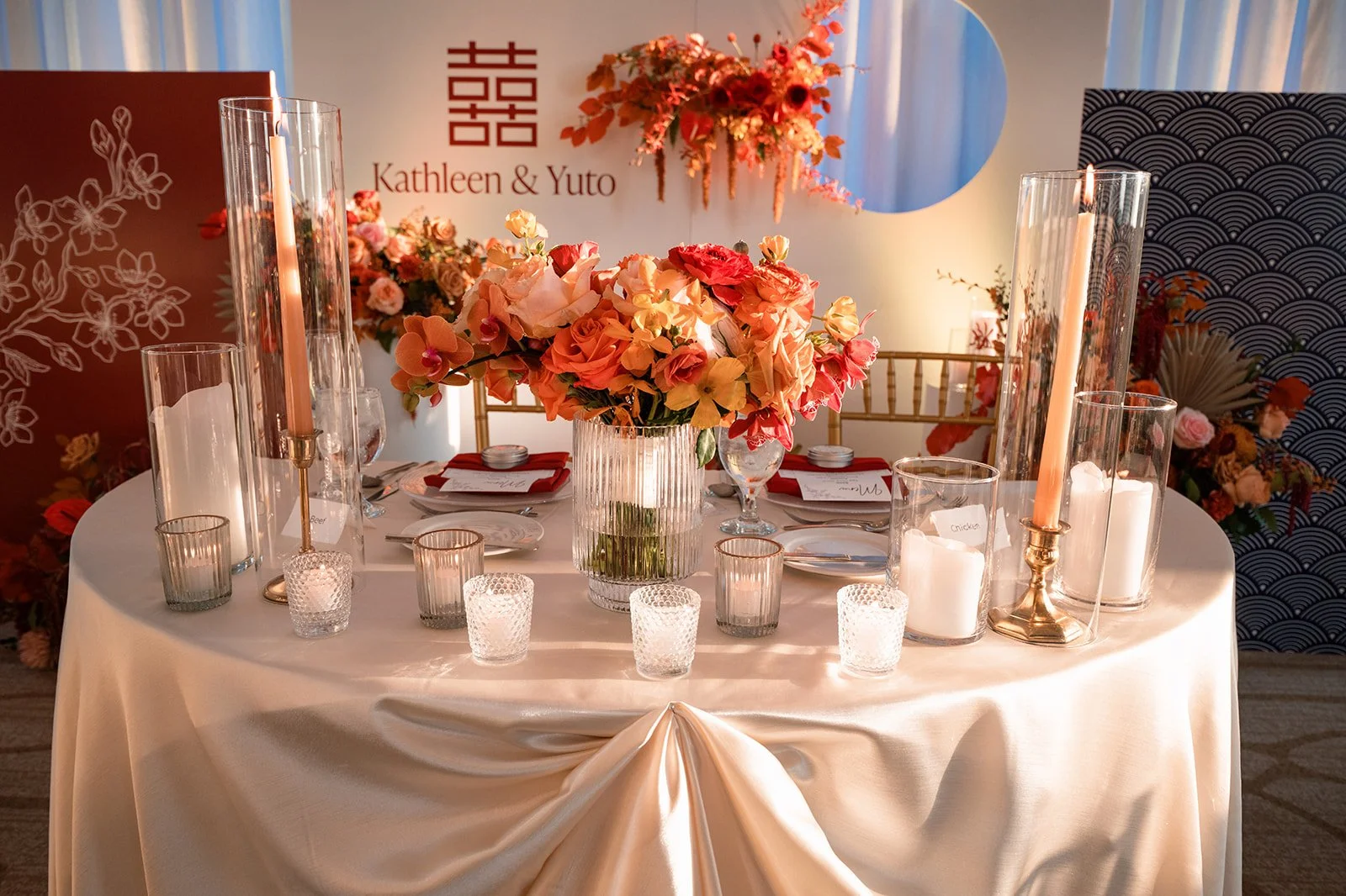 Wedding reception table with floral centerpiece, candles in glass holders, and elegant table settings, with a decorated backdrop featuring the names Kathleen and Yuto.