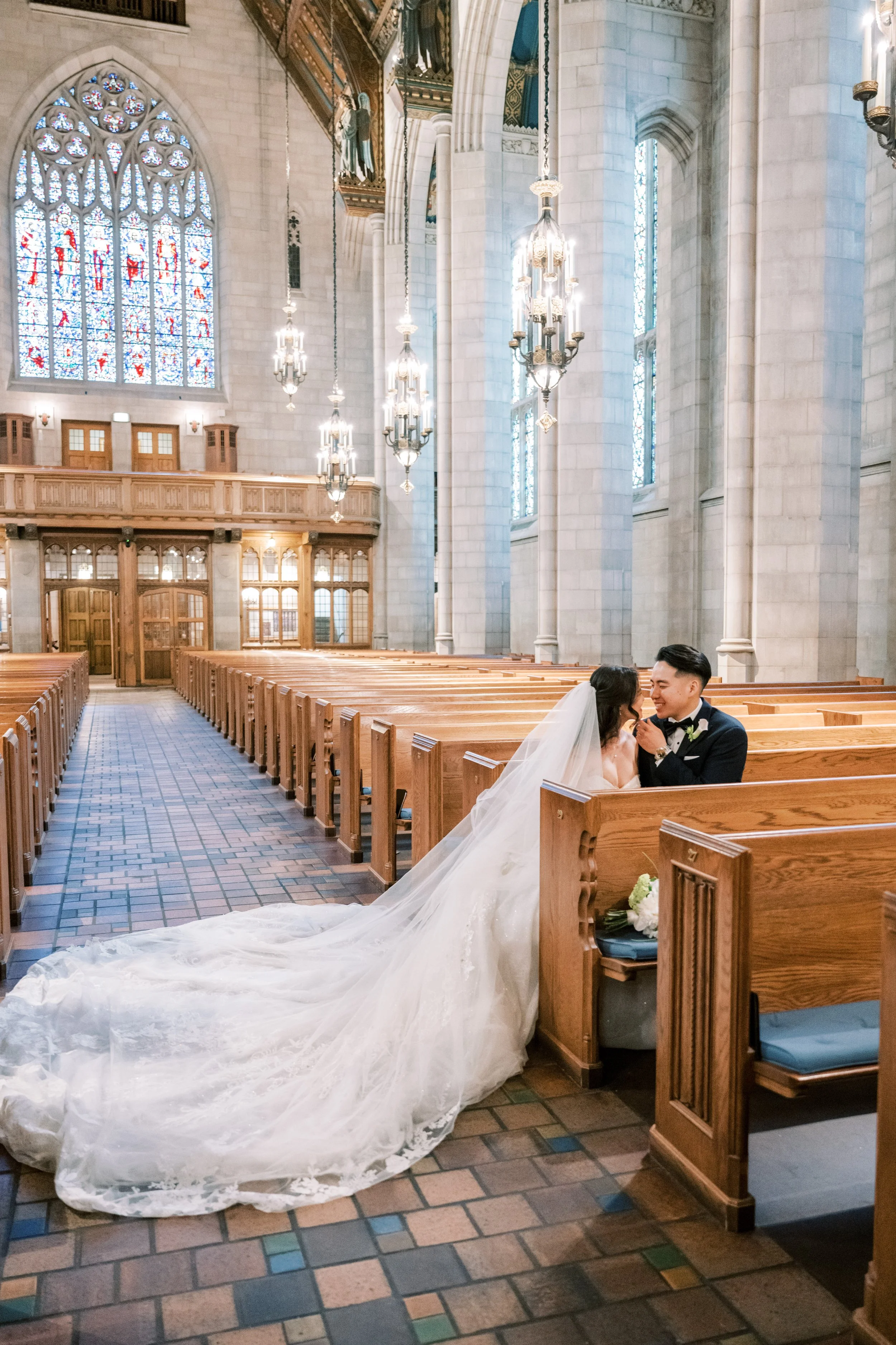 A bride and groom sitting in a church, sharing a joyful moment. The church interior features tall stained glass windows, ornate chandeliers, and wooden pews.