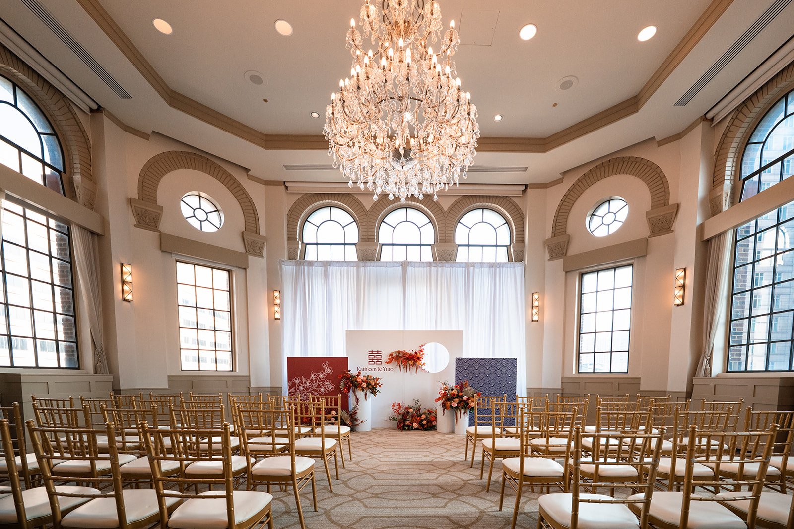 Elegant wedding ceremony setup in a bright, spacious room with a chandelier, large windows, and gold chairs arranged in rows facing a decorated backdrop with flowers.