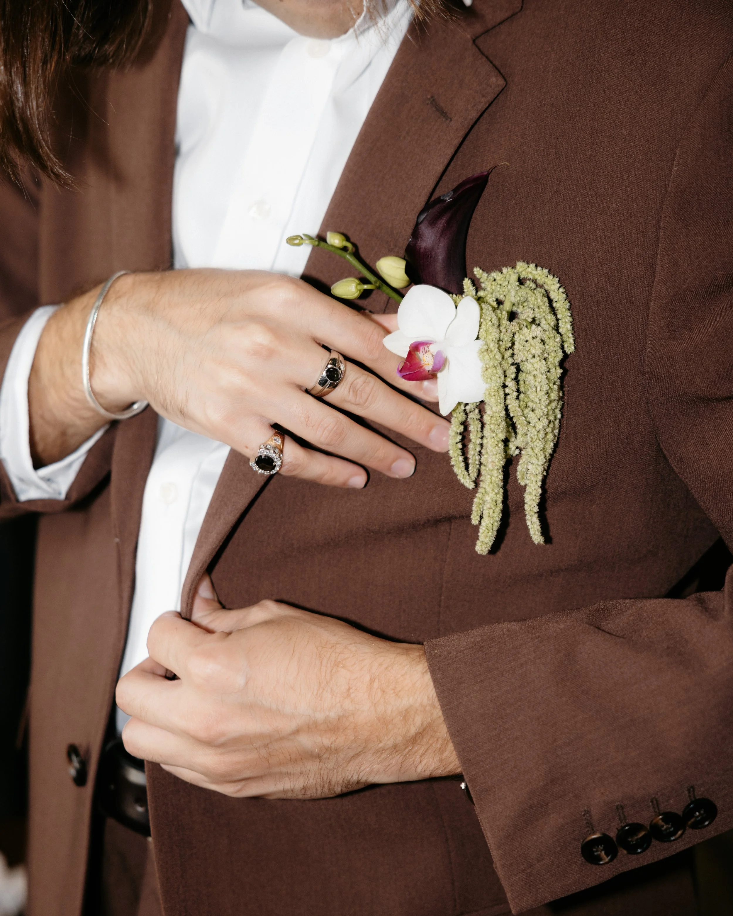 Close-up of a man in a brown suit with a white shirt and a boutonniere on the lapel. The boutonniere features a white orchid, dark purple calla lily, and various small filler flowers. The man's hands are adjusting his suit, and he is wearing rings an